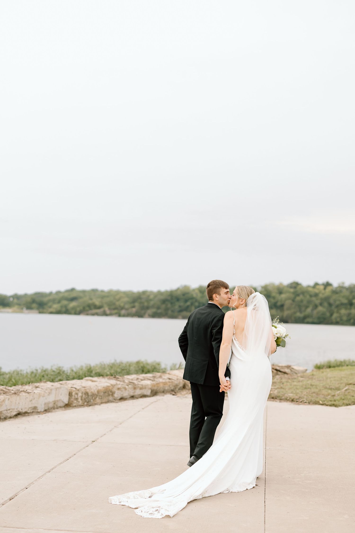 A romantic couple in wedding attire share an intimate moment on a lakeside pathway during their special day.