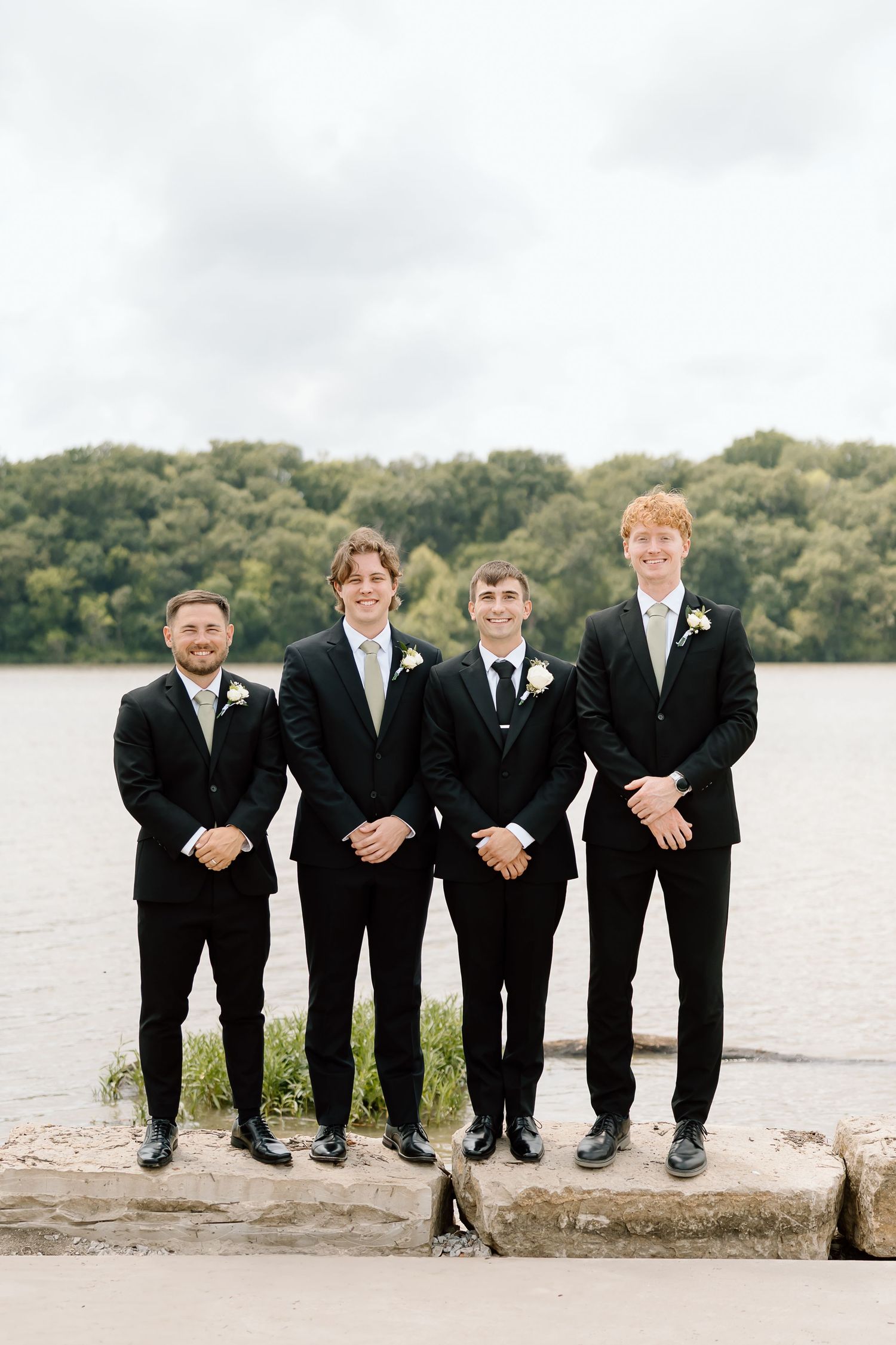 A groomsmen party poses sitting in a row on a stone ledge near a tranquil lake in black suits.