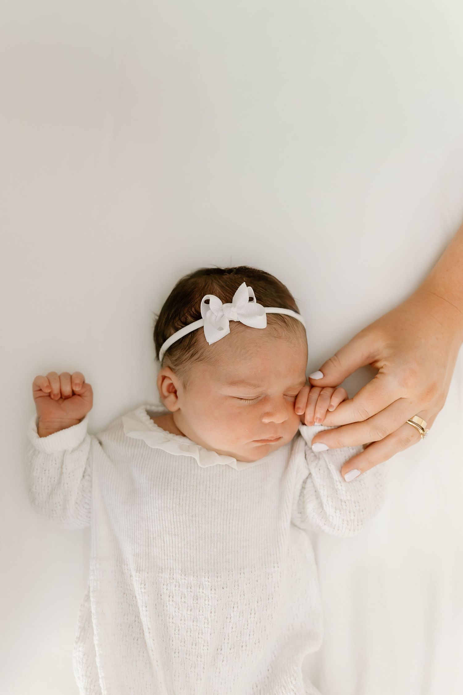A sleeping newborn baby wearing white lies on a light background while gentle hands touch their head and arms.