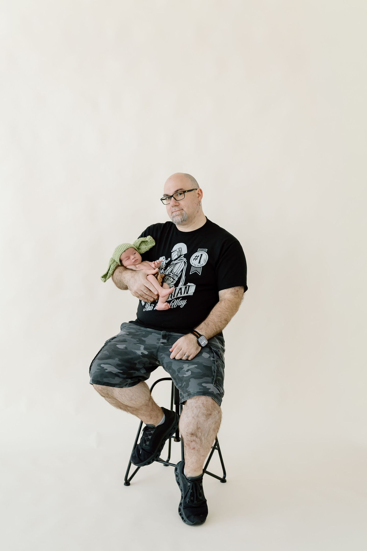 Person in black t-shirt and camo shorts sits on chair with green plant in minimalist white studio setting.