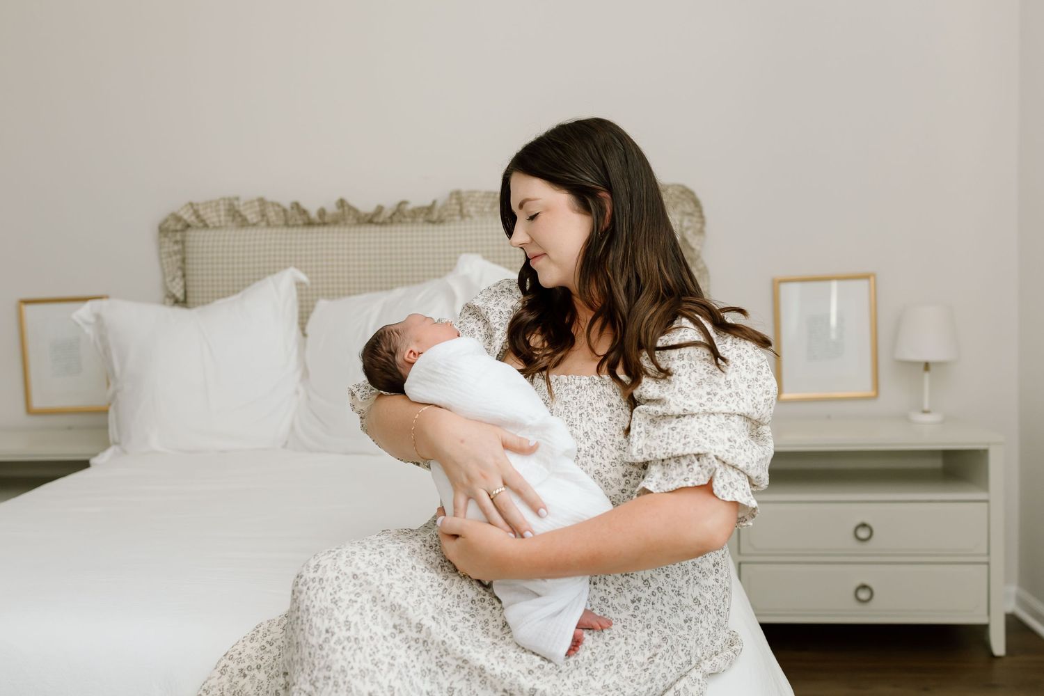 A mother and newborn baby share tender moments together on a white bed in a softly lit bedroom.