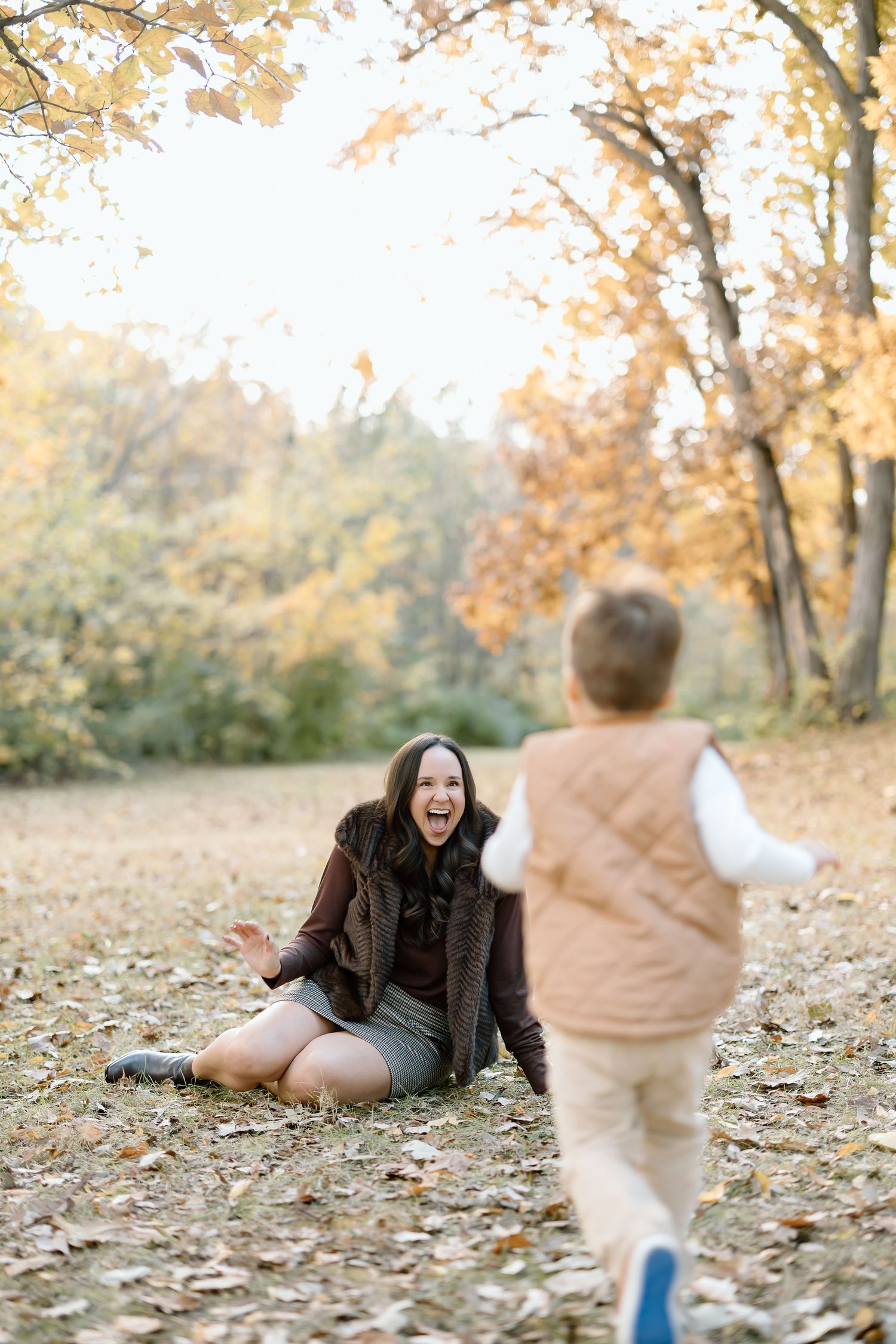 A joyful autumn scene with someone sitting on fallen leaves while a child in a vest runs towards them in a park.
