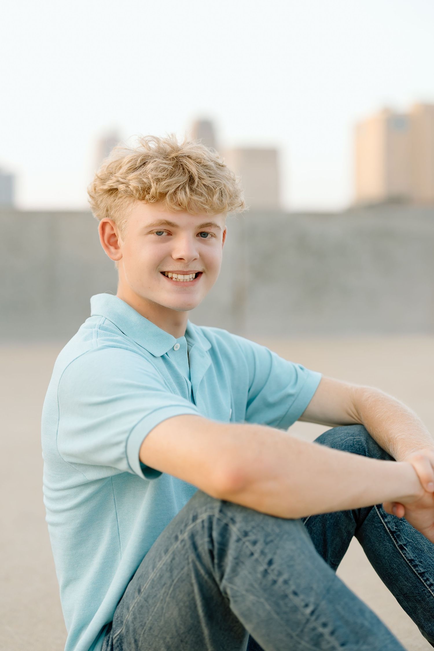 Young person in light blue shirt and jeans sits casually outdoors with cityscape in background.