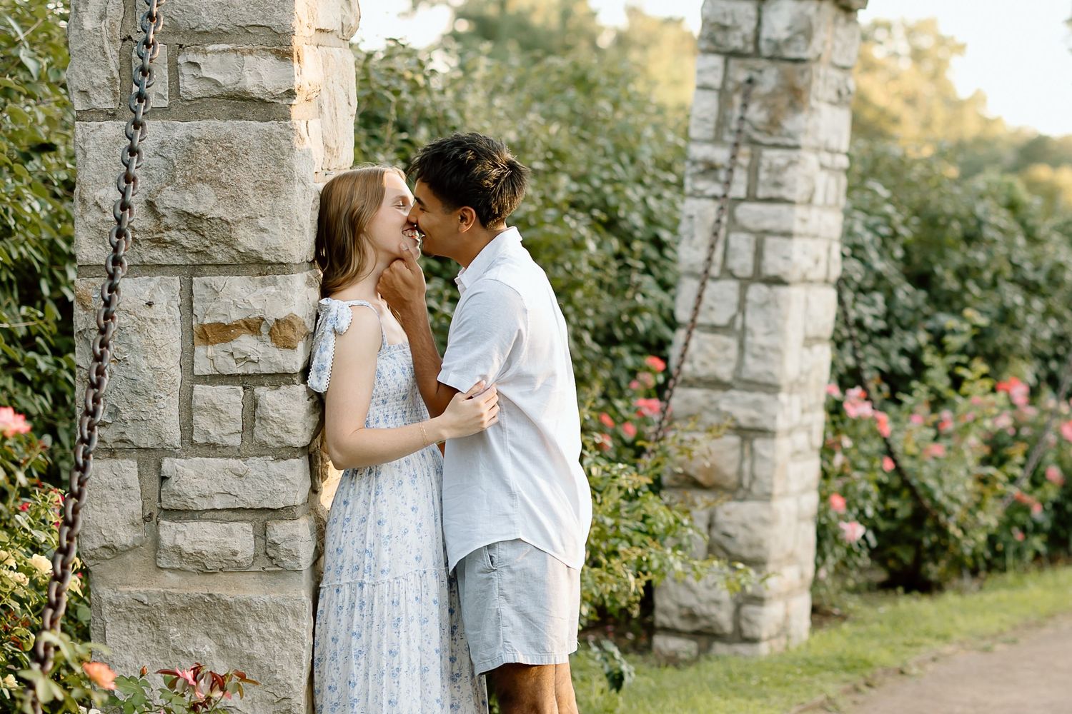 A romantic moment between two people sharing a tender kiss by stone pillars in a garden setting at sunset.
