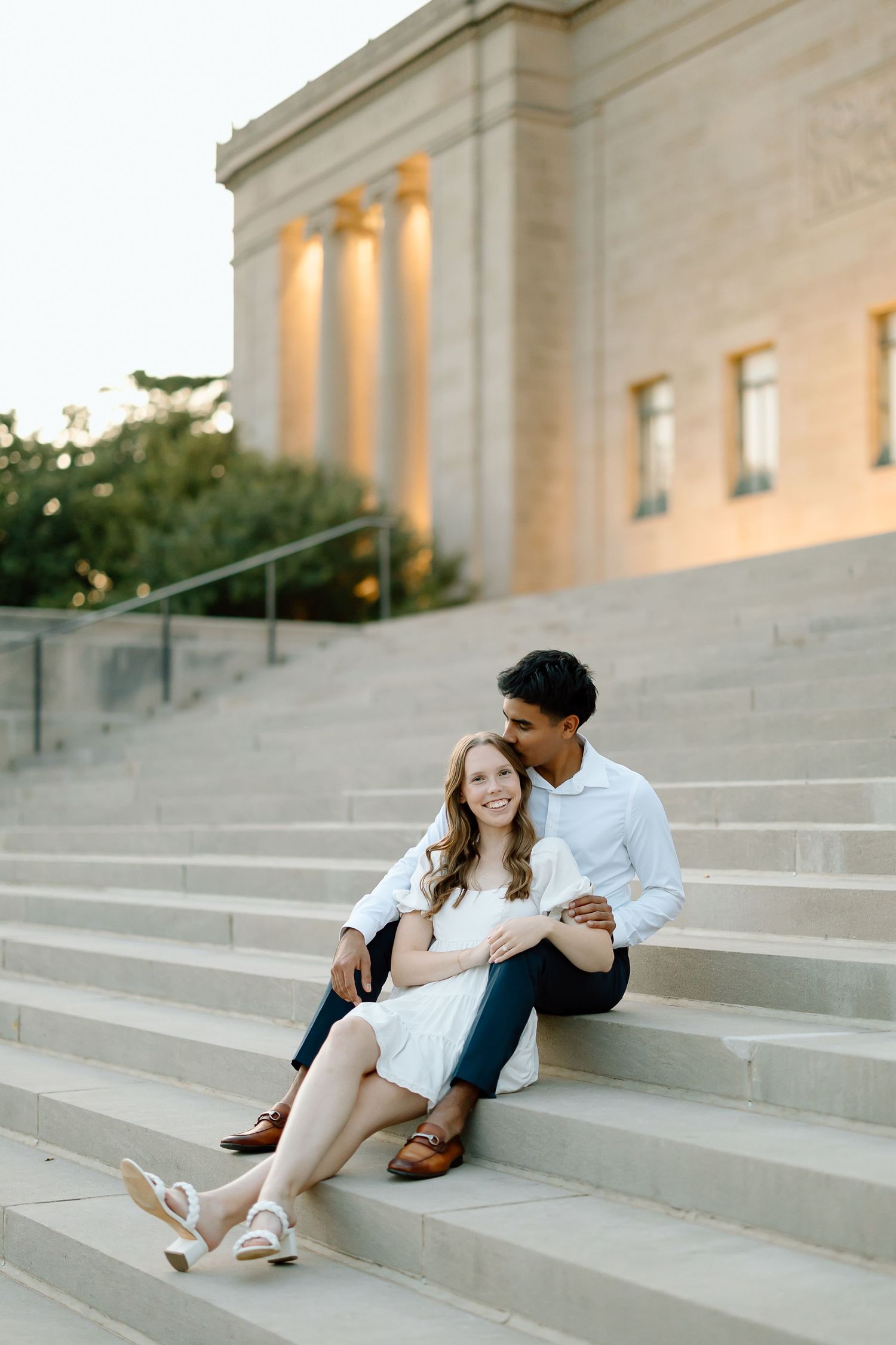 A couple sits intimately on concrete steps at sunset with warm lighting on a classical building in the background.