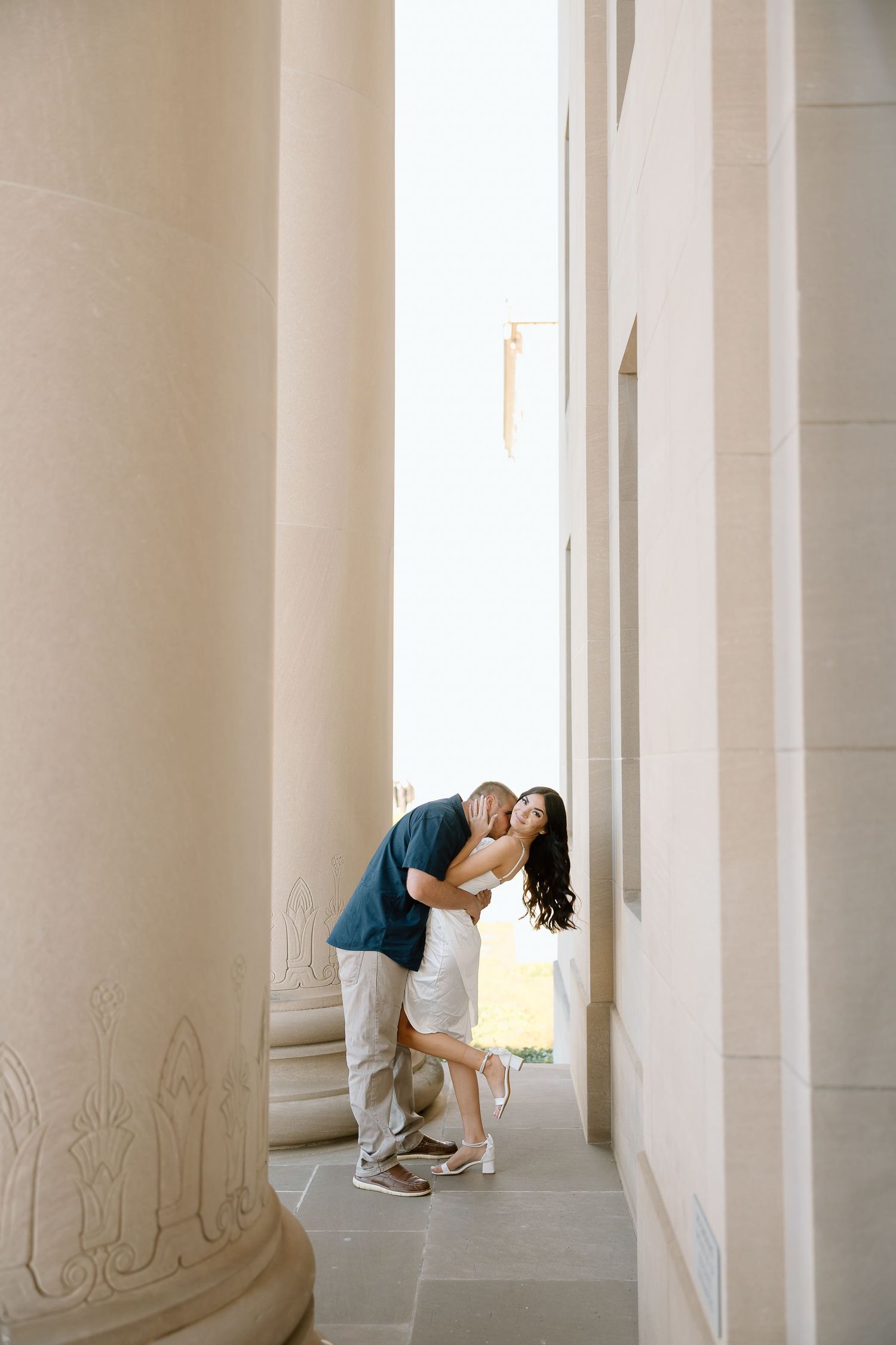 A couple embraces in a romantic kiss while standing in an elegant white colonnade hallway.