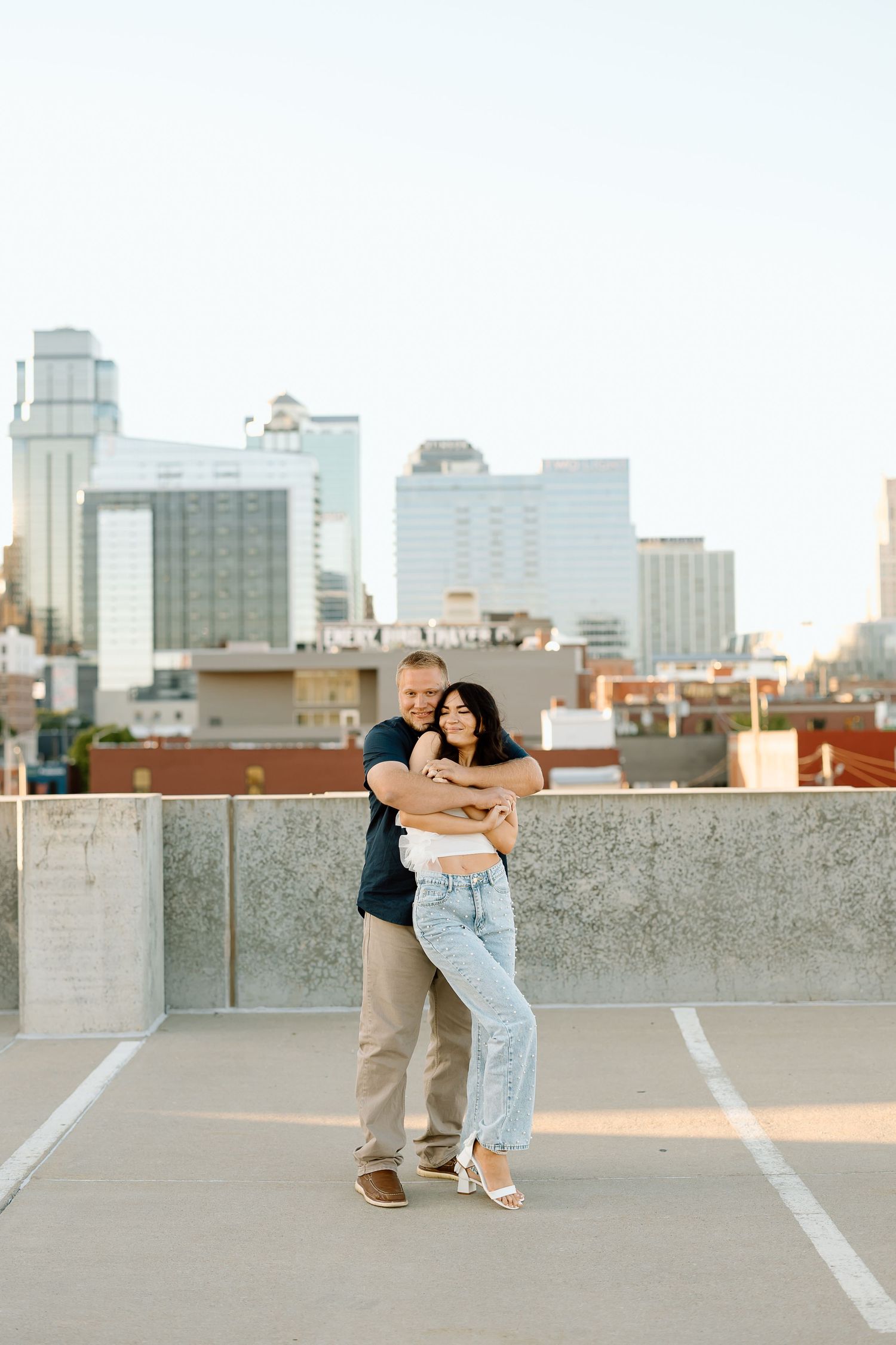 A couple embraces on a rooftop parking garage with a city skyline in the background during sunset.