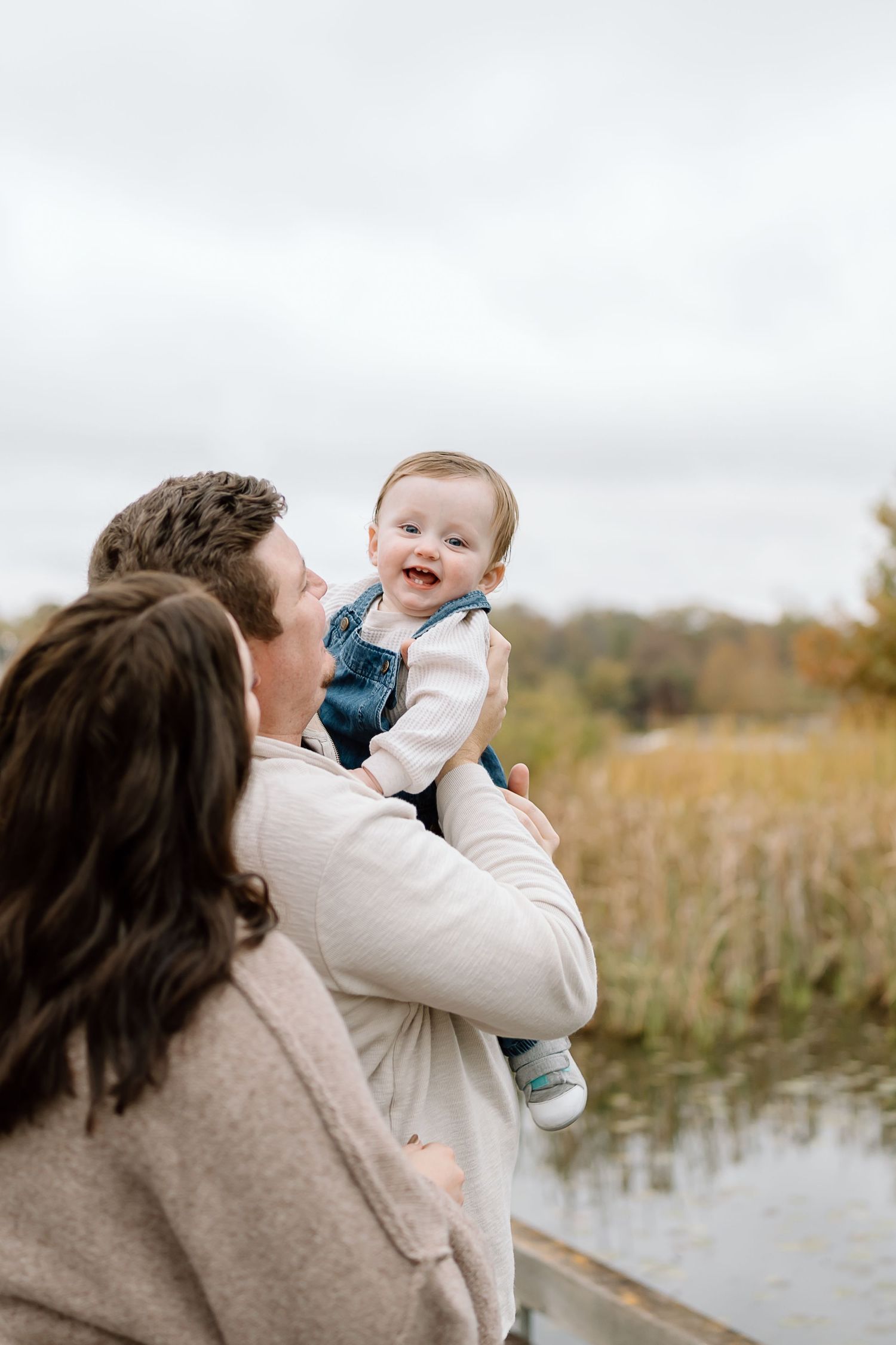 Parent lifts and embraces young baby wearing blue outfit in autumn field near water during outdoor photo session.
