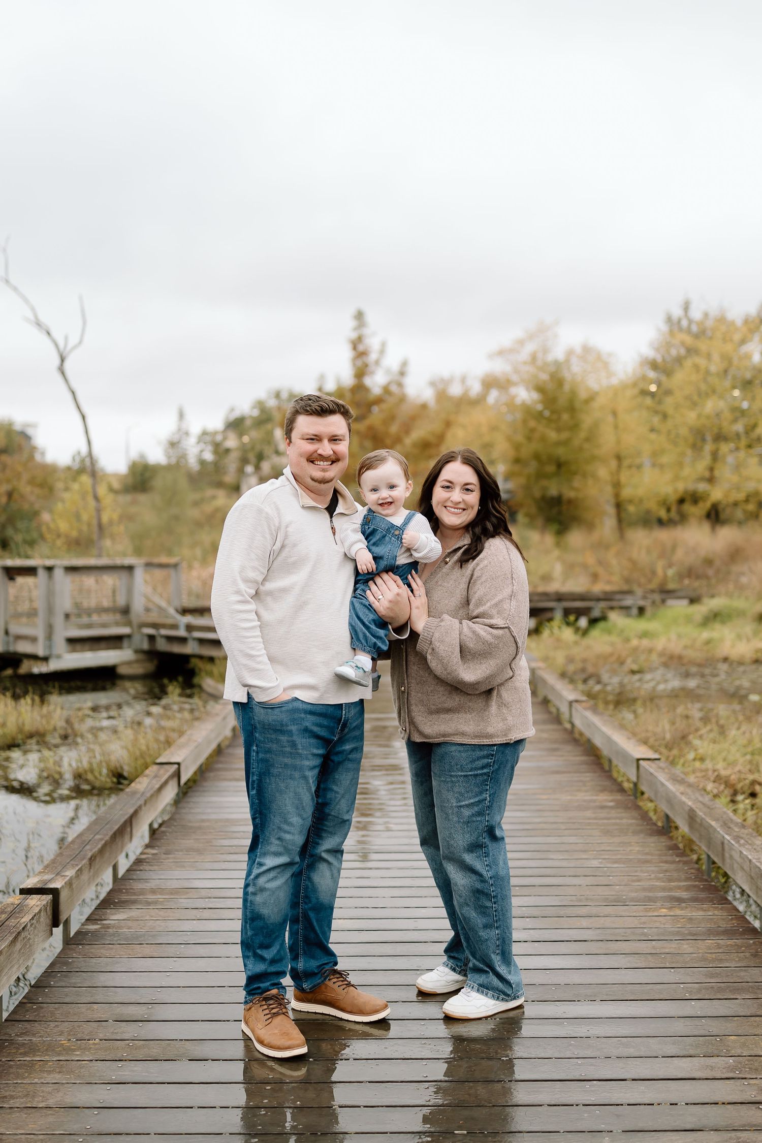 A family stands together on a wooden boardwalk surrounded by autumn foliage in a natural outdoor setting.