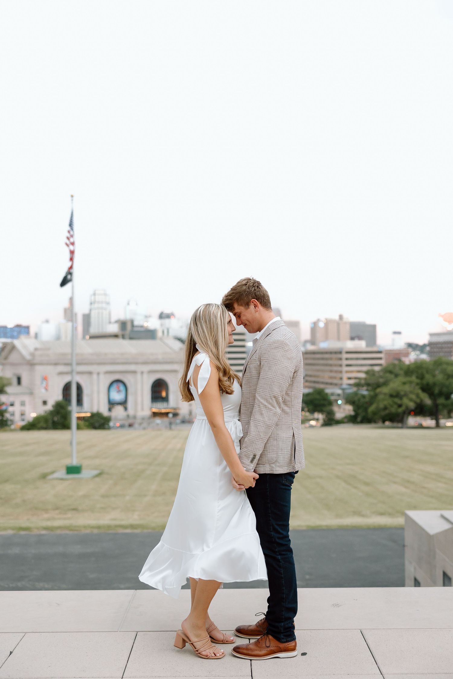 A couple in wedding attire shares romantic kisses while standing on a paved outdoor area with city views.