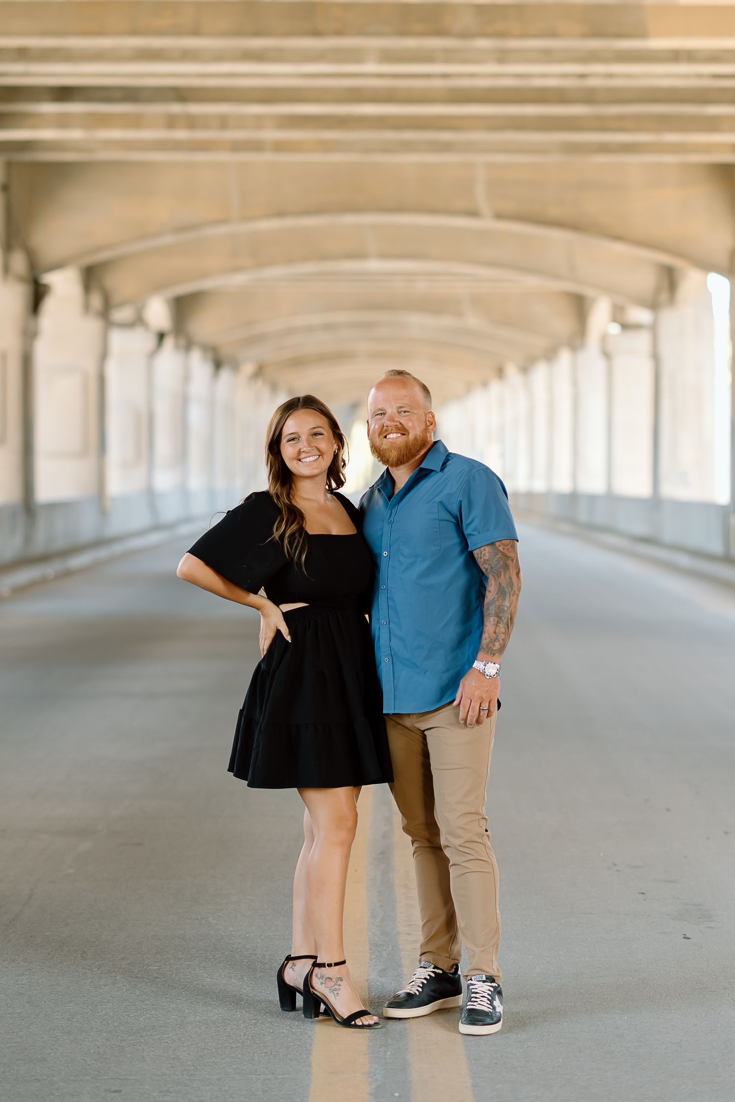 A couple poses playfully in a covered walkway with concrete columns and natural lighting.