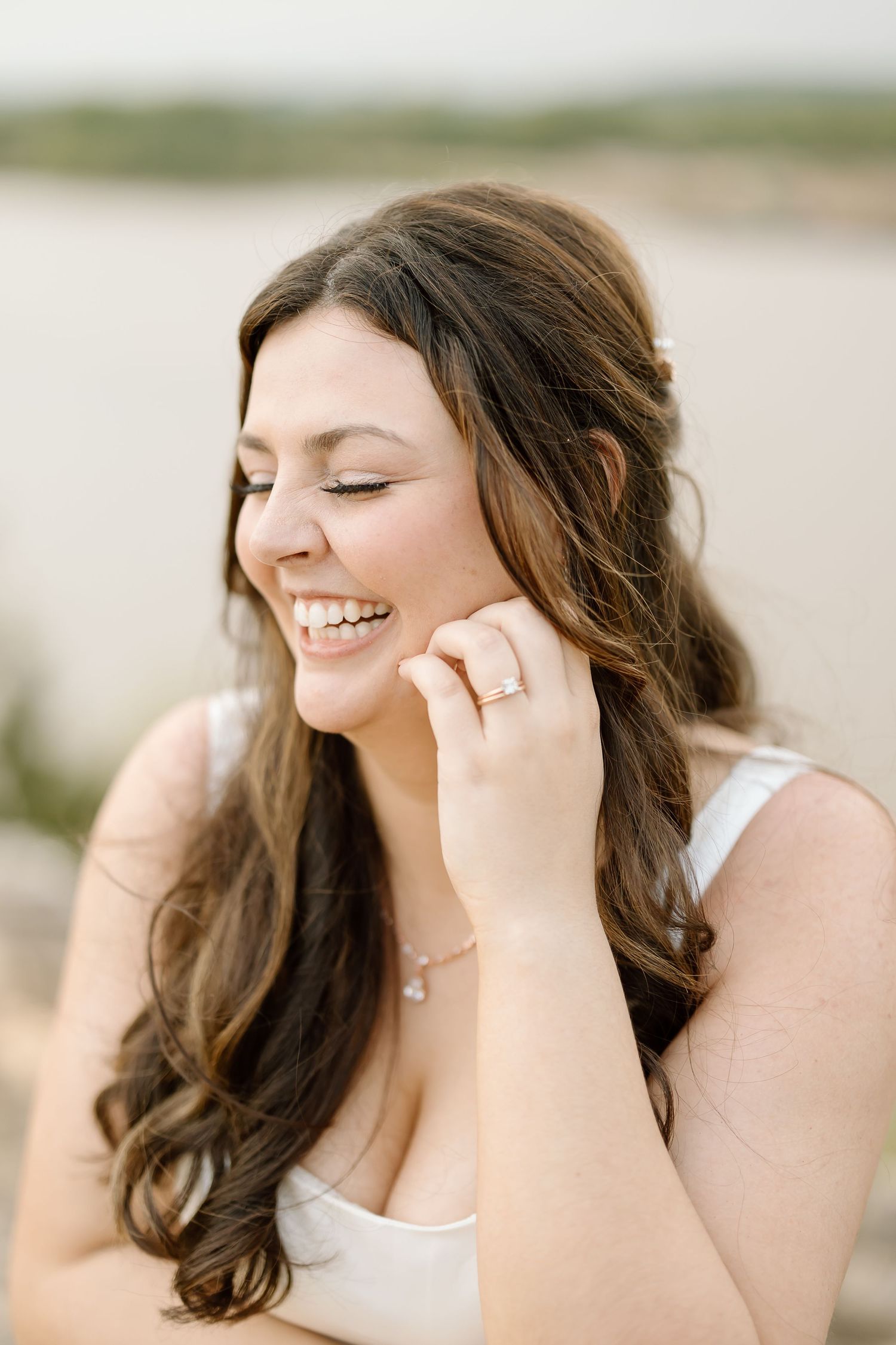 A person with long brown hair laughs joyfully while covering their mouth with their hand in an outdoor setting.