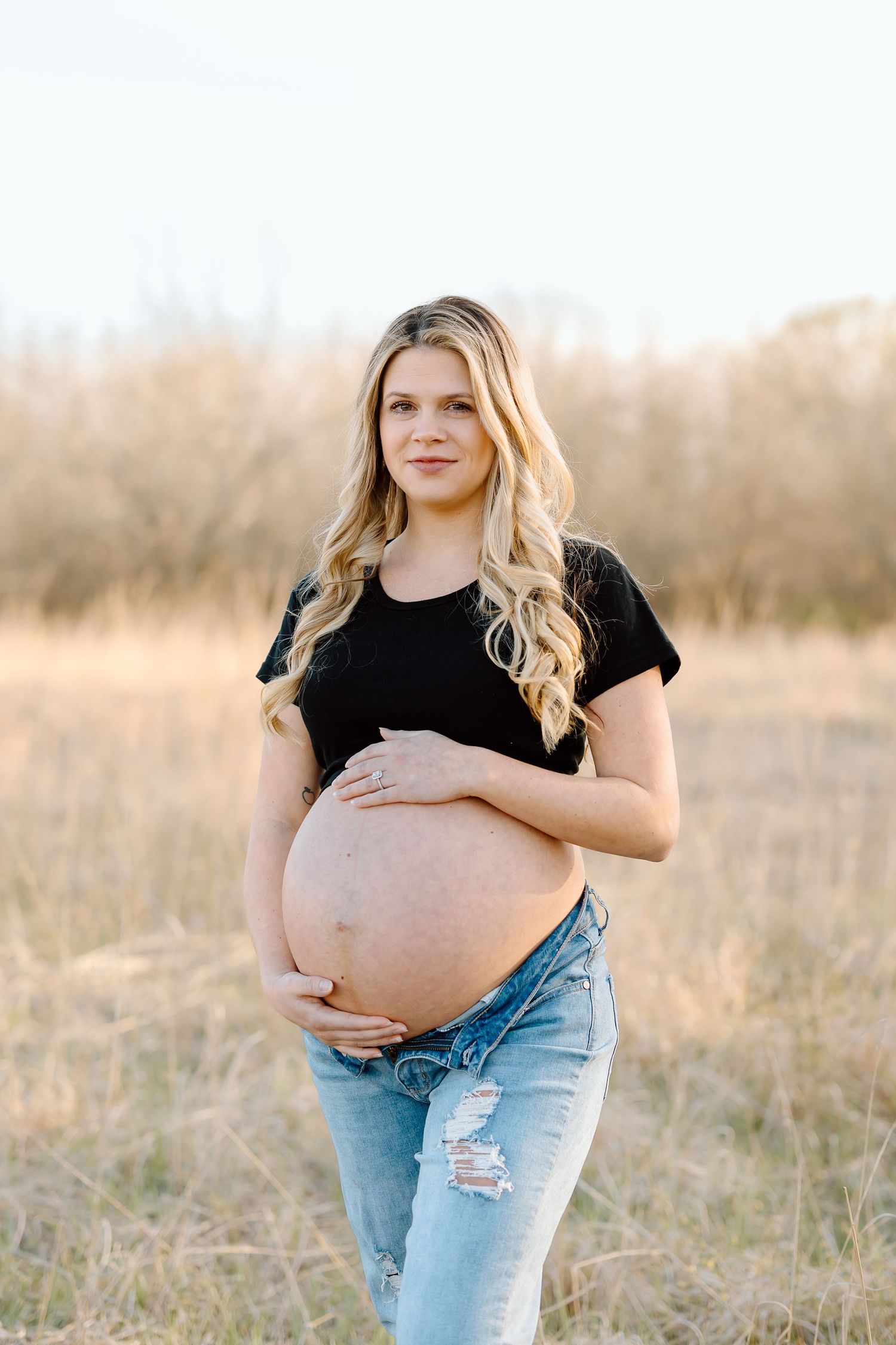 A maternity portrait session features someone in casual attire posing in tall grass during golden hour.