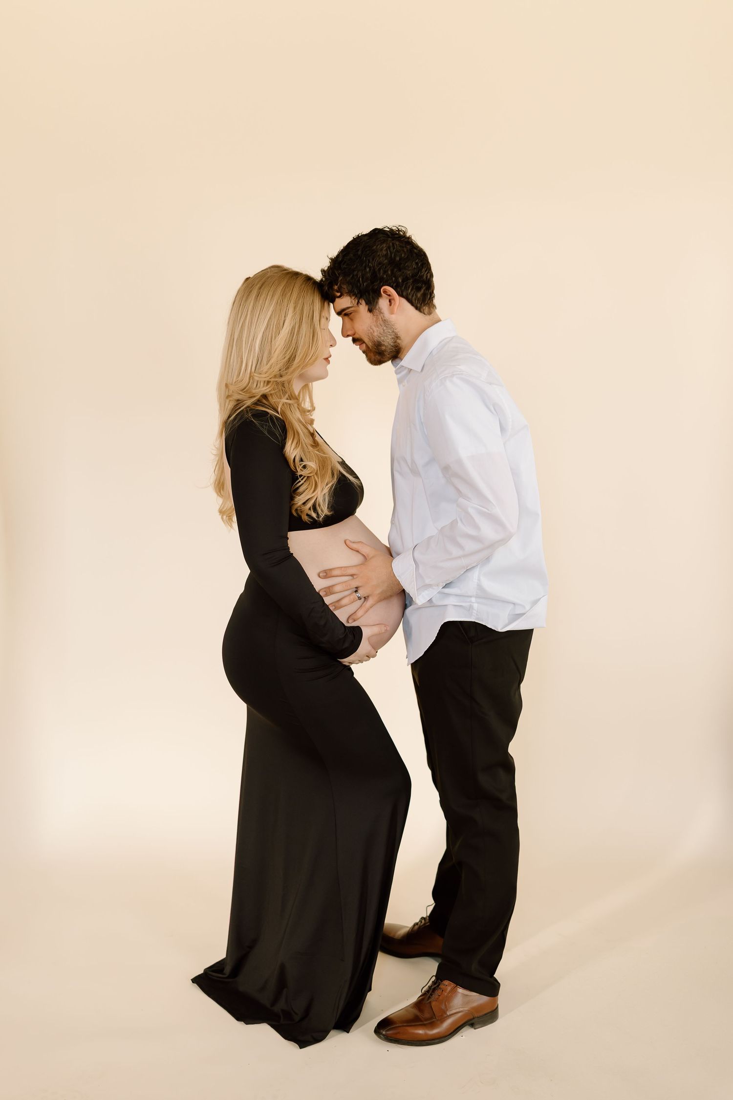 Intimate maternity portrait with couple in tender embrace against beige backdrop.