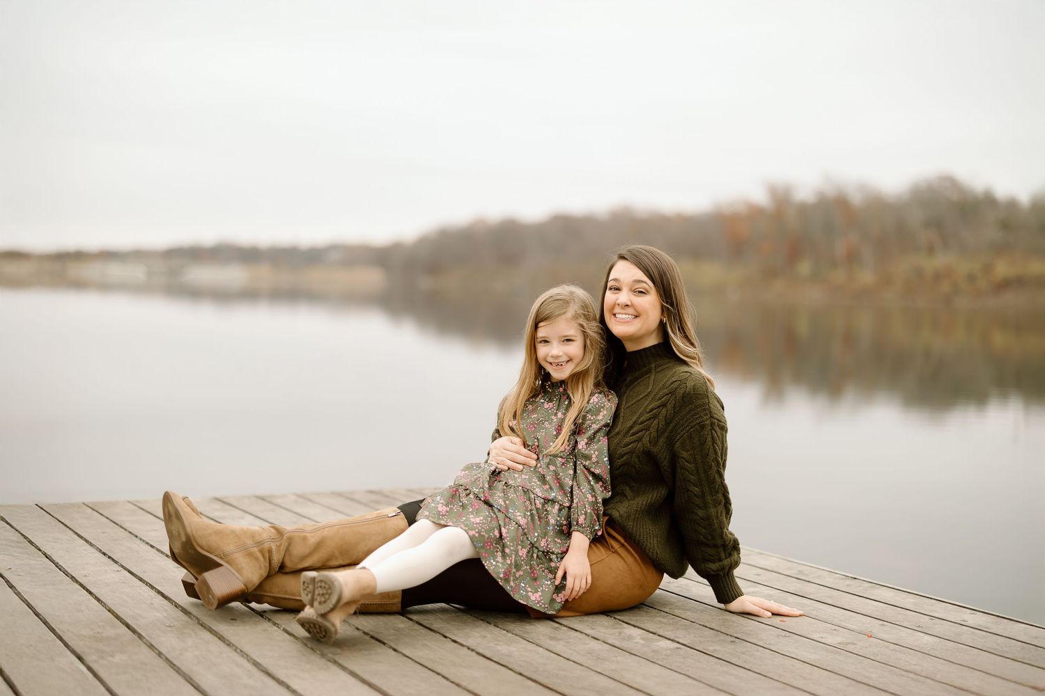 Parent and child share relaxed moment sitting together on wooden dock by peaceful lake.