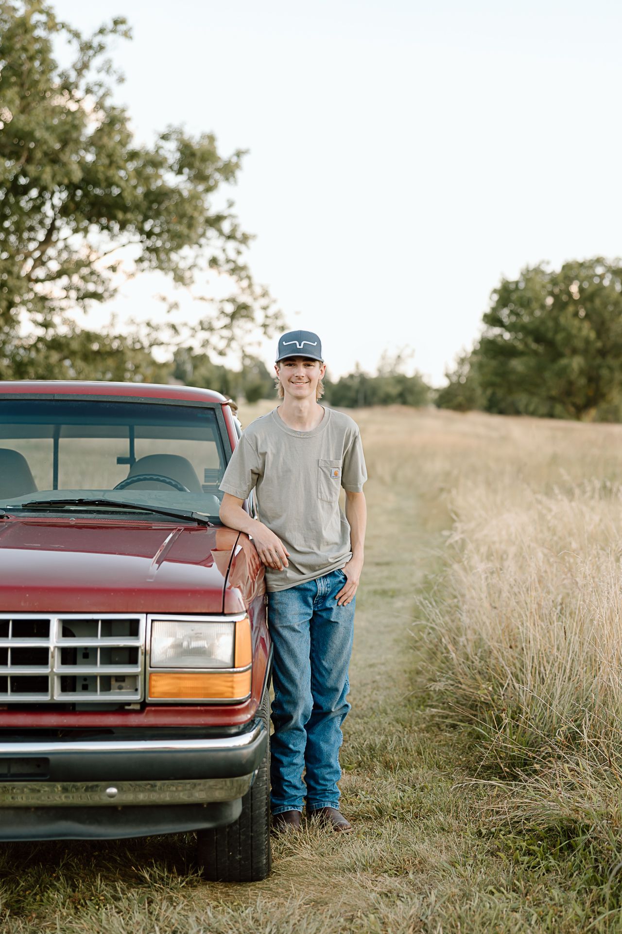 Someone in casual attire standing next to a classic red Ford pickup truck in a grassy rural setting.