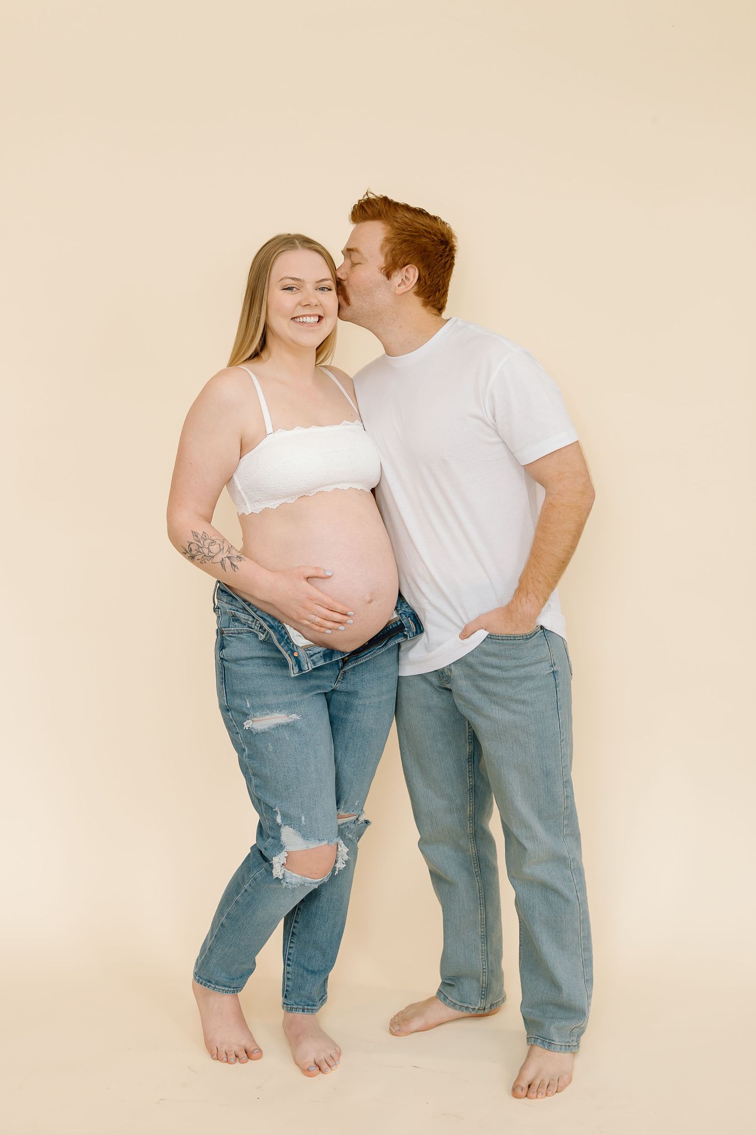 Expectant couple in casual attire share an intimate moment against beige wall during maternity photoshoot.