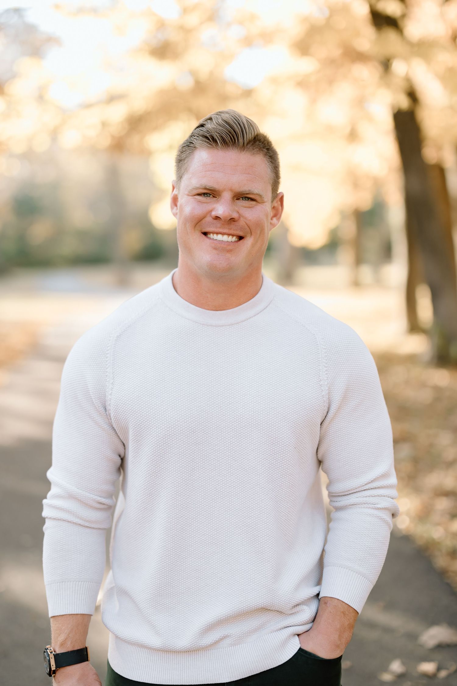 Portrait of a man in a white sweater standing outdoors with autumn trees behind him.