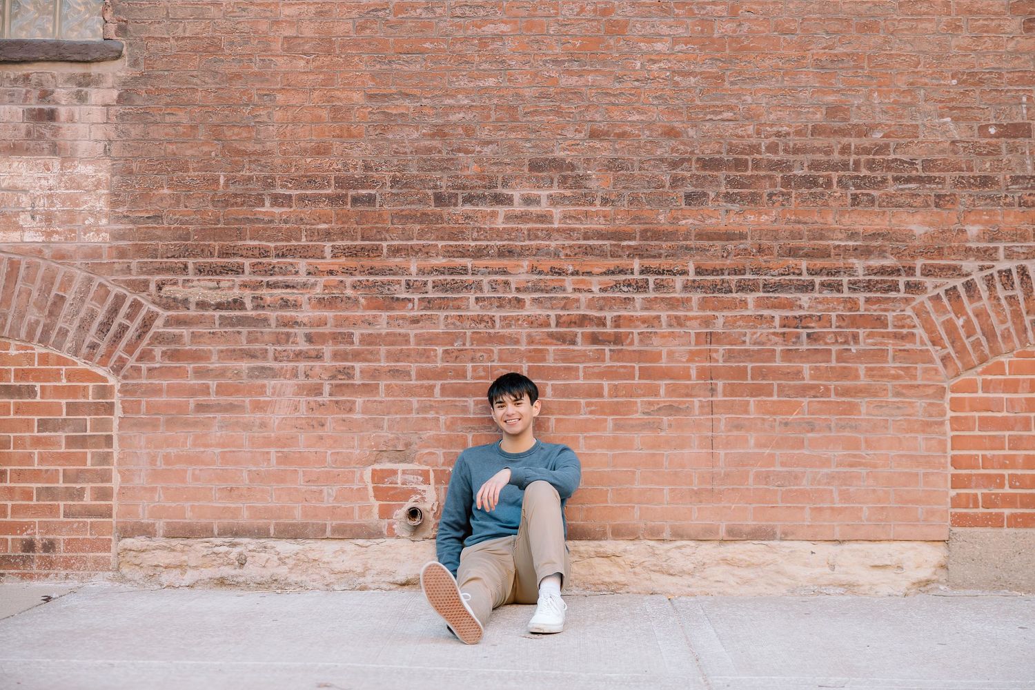 Person in casual attire sitting against red brick wall holding beverage in urban setting.