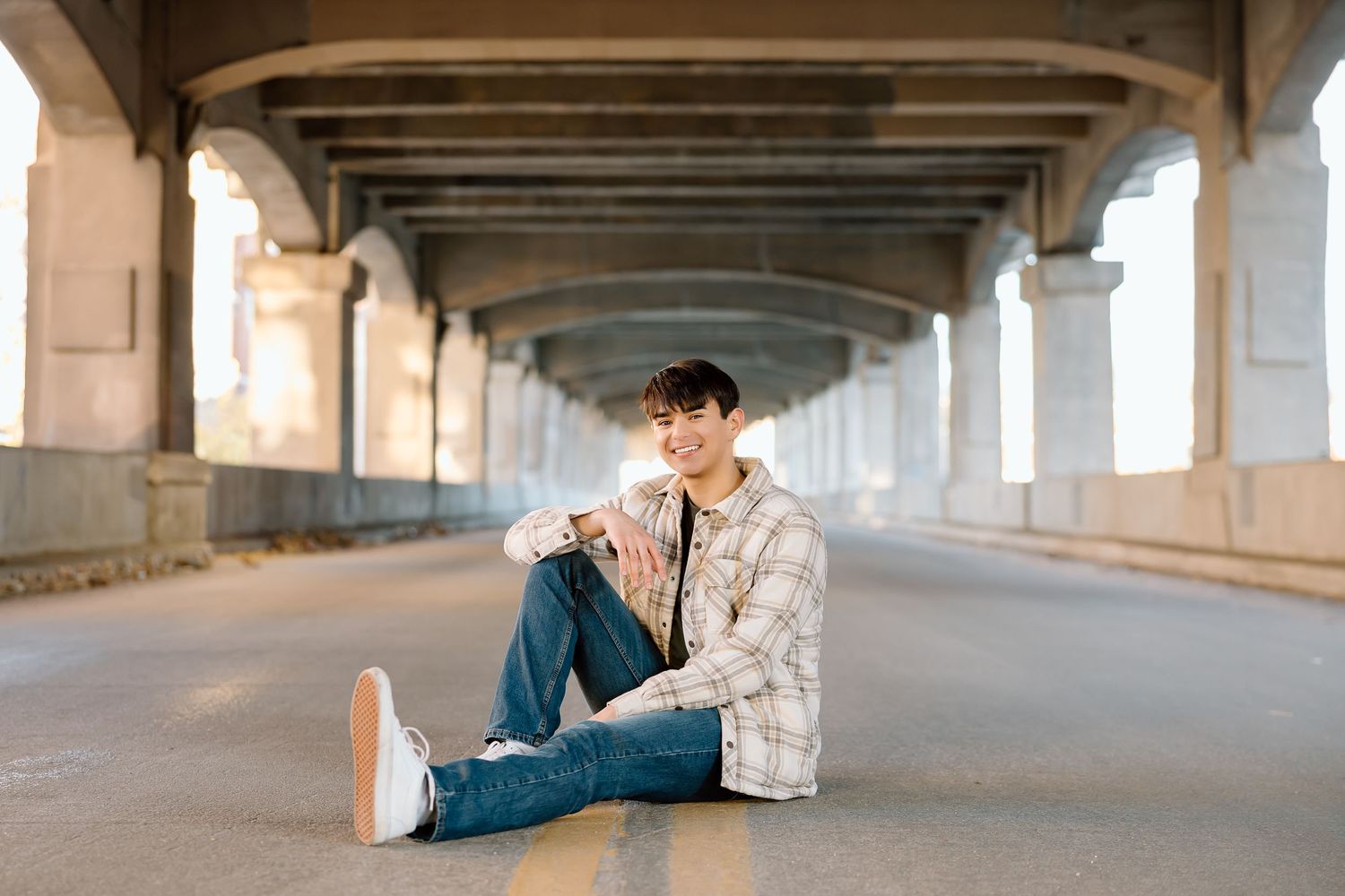 Relaxed pose on ground under bridge wearing light plaid jacket and jeans.