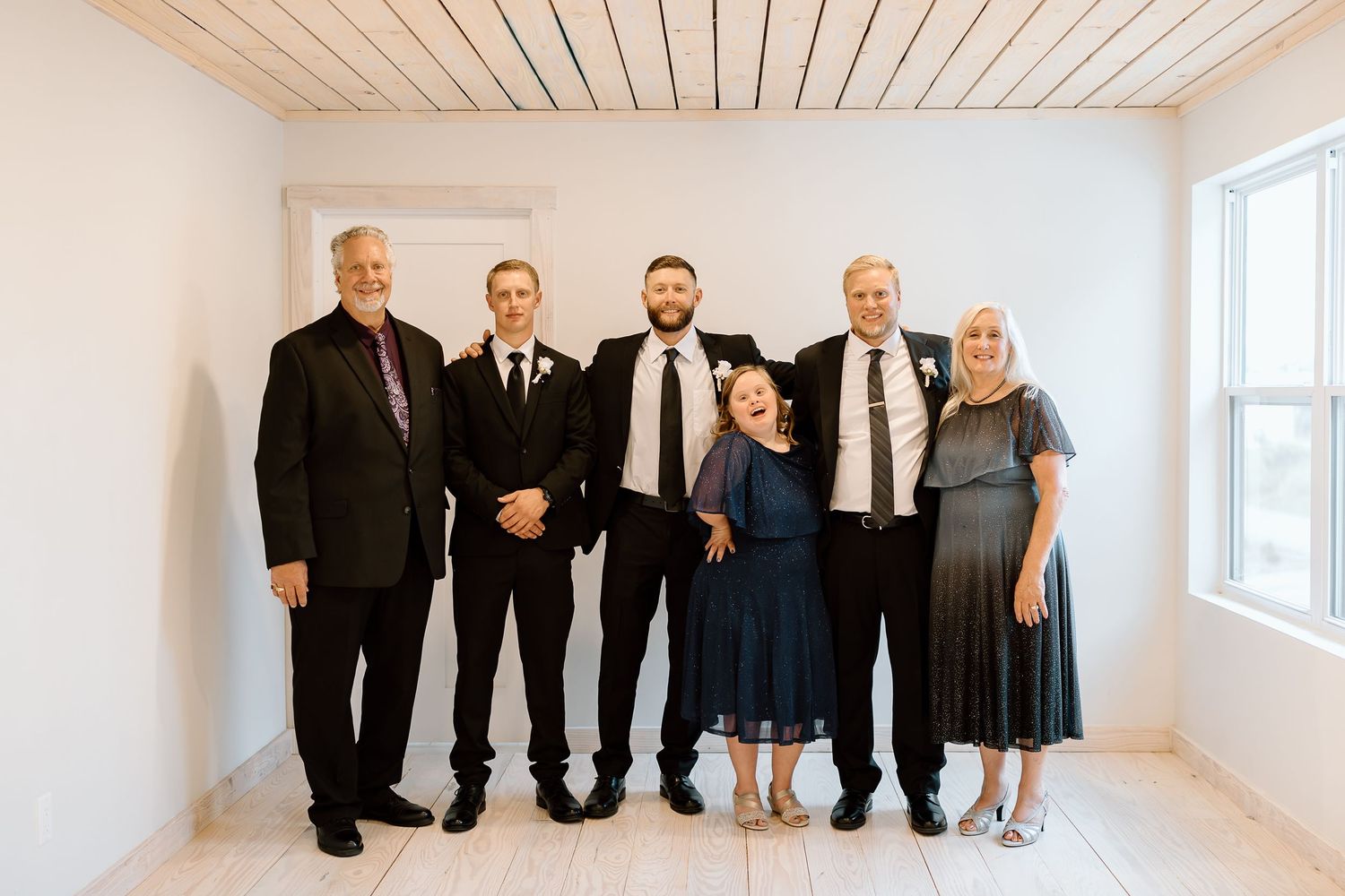 A wedding party stands in a row against a white wall with wooden ceiling panels and bright natural lighting.