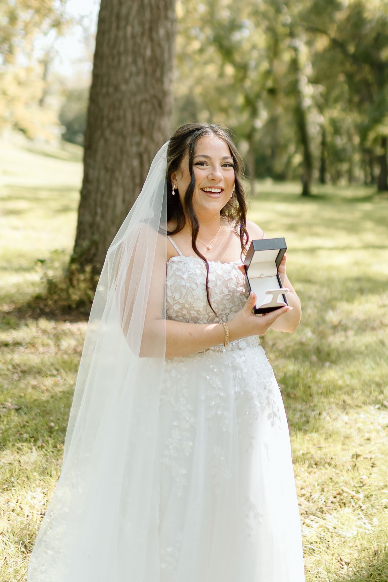 A bride in a white lace wedding dress laughs joyfully while holding a small box in an outdoor garden setting.