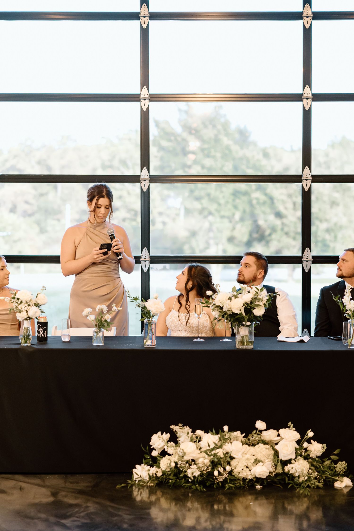 Wedding party seated at head table with elegant white floral arrangements during reception speeches.
