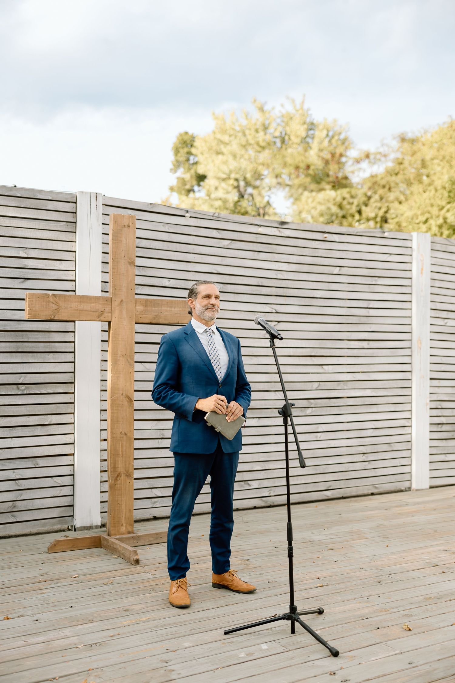 A person in a blue suit kneels on a wooden deck next to a wooden cross during an outdoor ceremony.