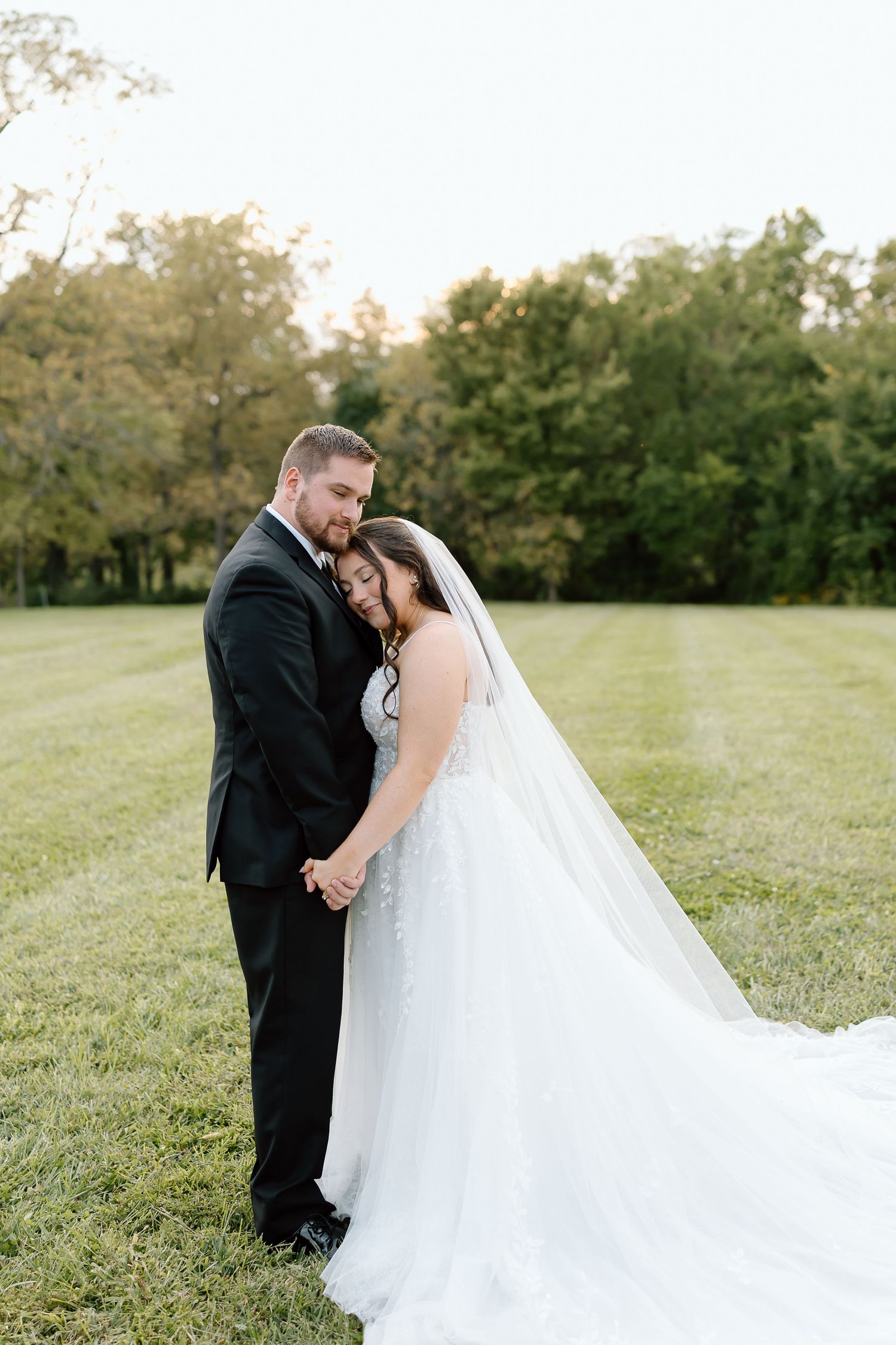Newlywed couple shares intimate moment in outdoor garden setting with flowing white wedding gown and black suit.