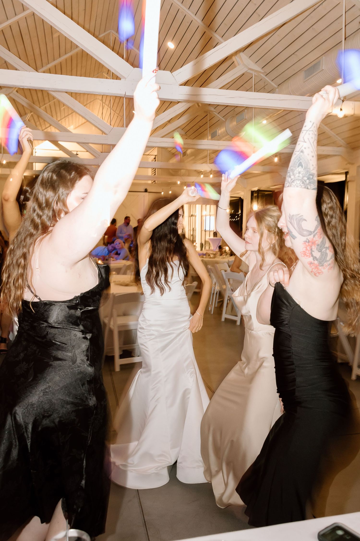 Wedding guests dancing and raising their hands on a dance floor under wooden beams and pendant lights.