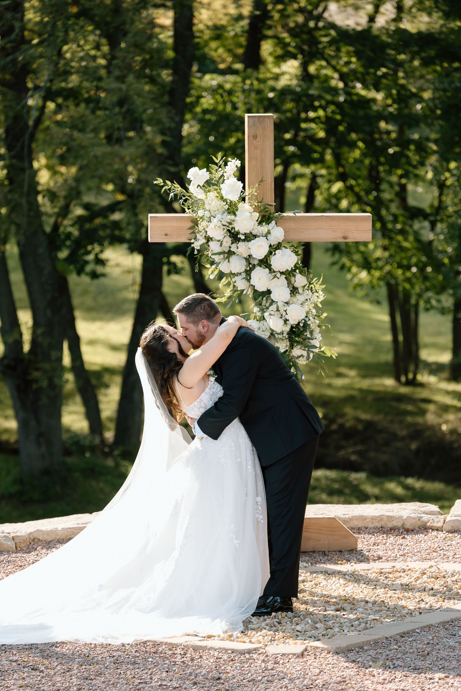 A couple shares a romantic kiss in front of a wooden cross decorated with white flowers in a sunlit outdoor setting.
