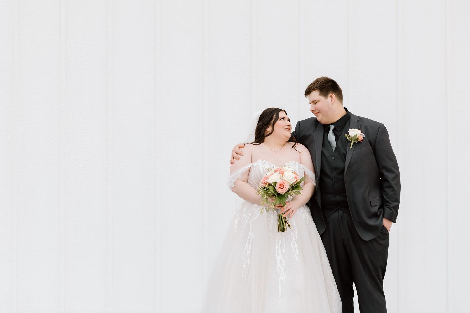 A couple in wedding attire stands together against a white wall, with the bride holding a pink and white bouquet.