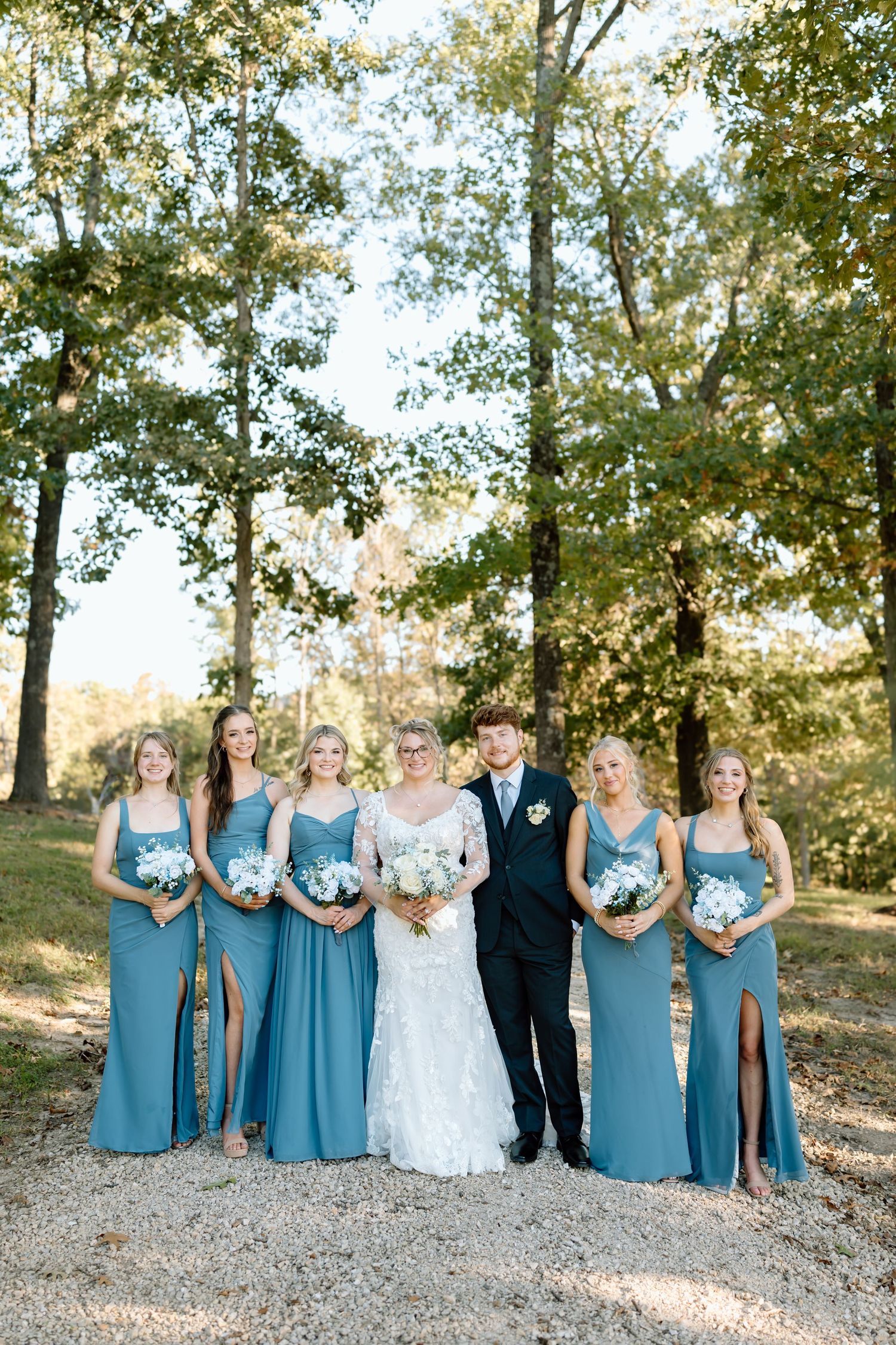 Wedding party in matching teal blue dresses and dark suits poses outdoors among autumn trees on a sunny day.