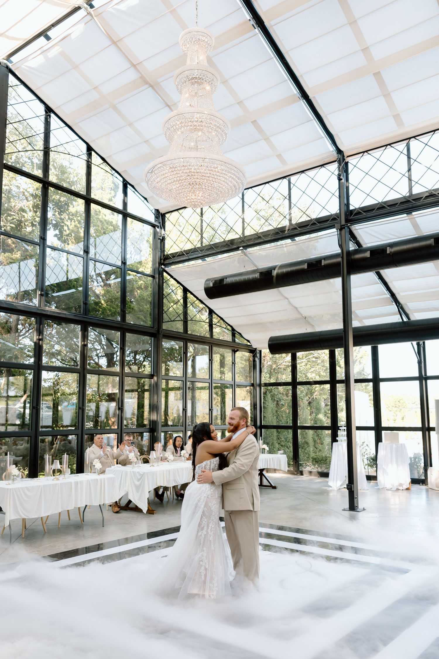 Bride and groom share first dance under crystal chandelier in elegant glass greenhouse wedding venue.