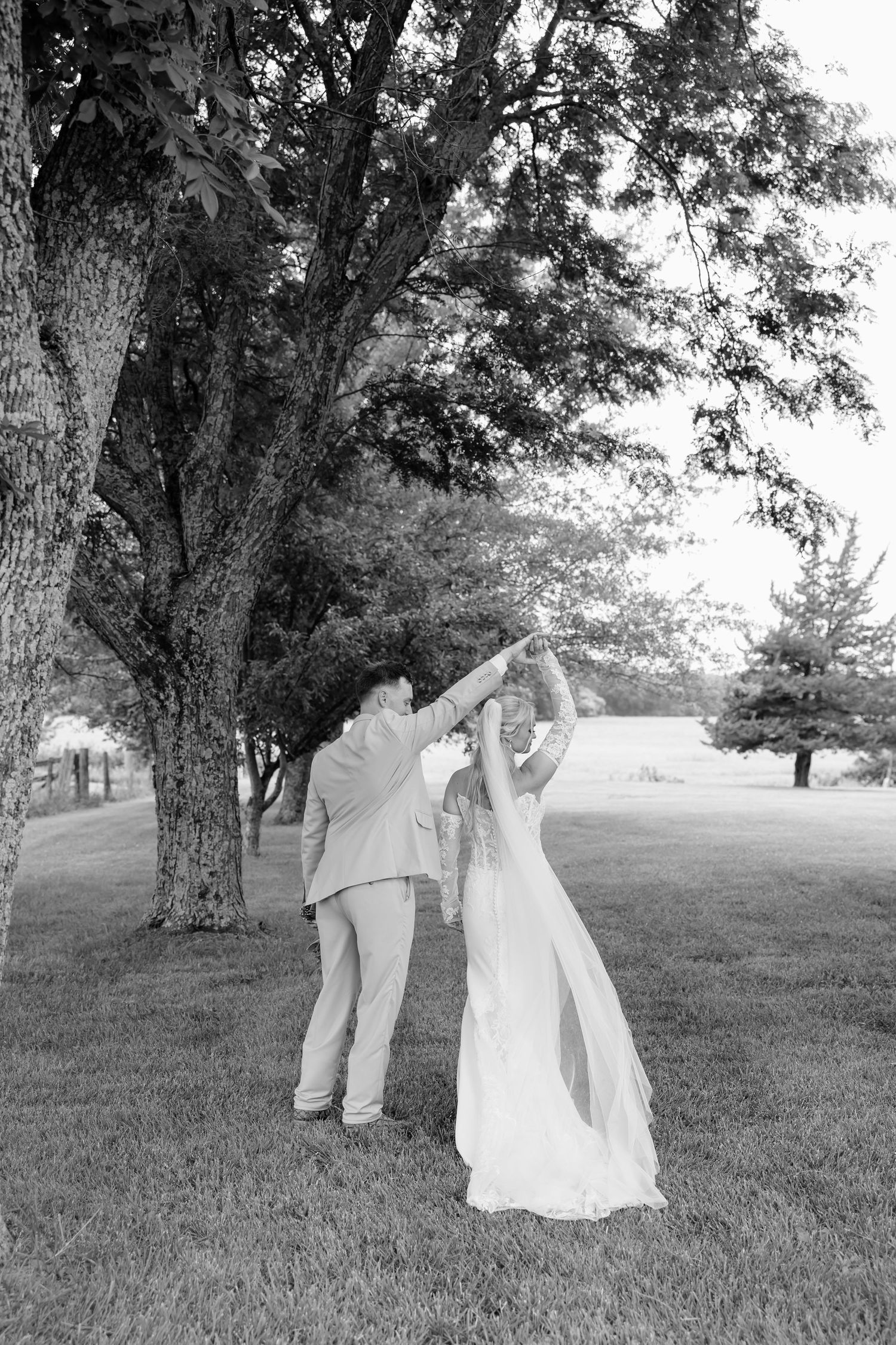 Black and white photo of a bride and groom dancing and spinning under a large tree on a green lawn.
