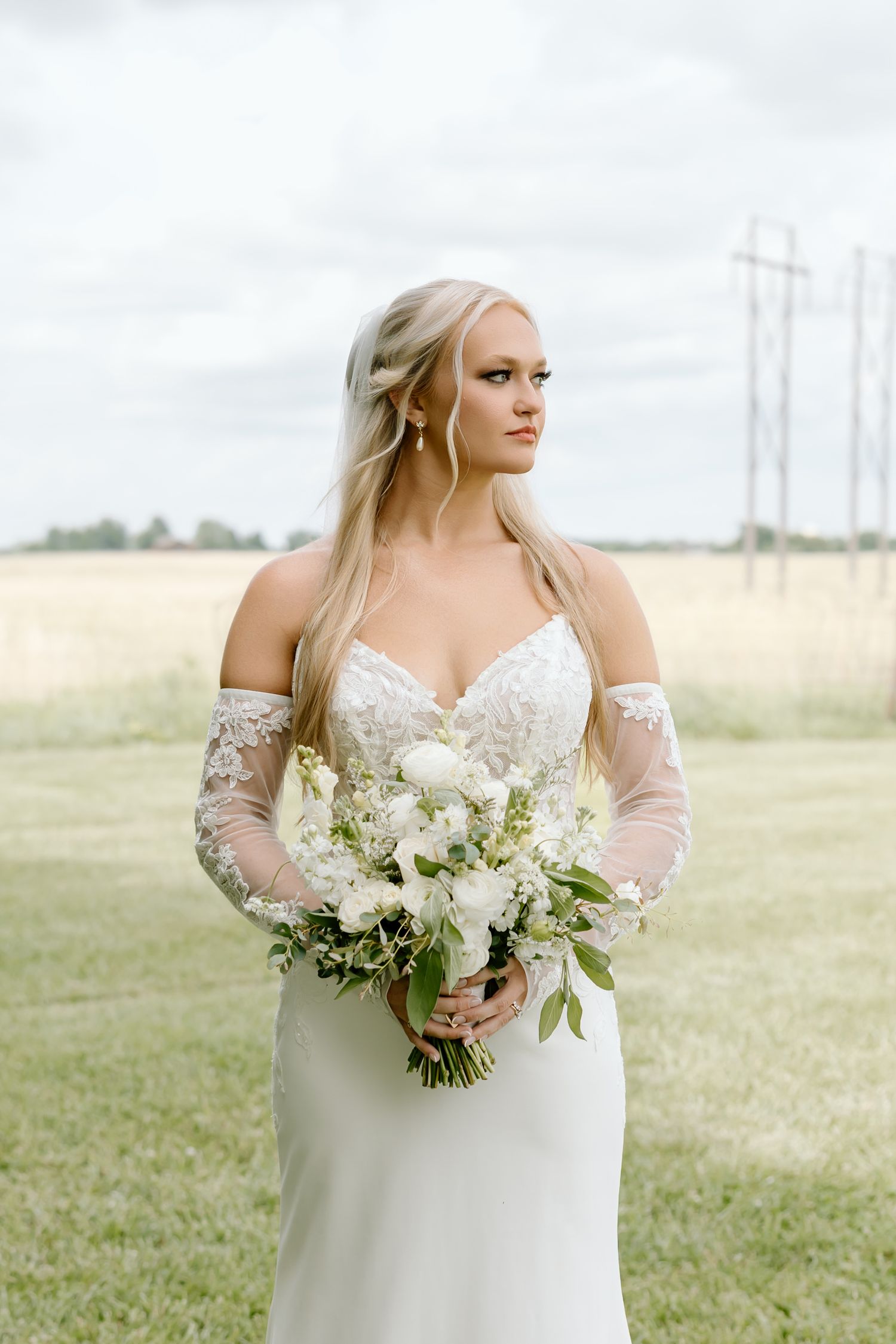 Blonde bride in off-shoulder lace gown holds white floral bouquet while standing in a green field.