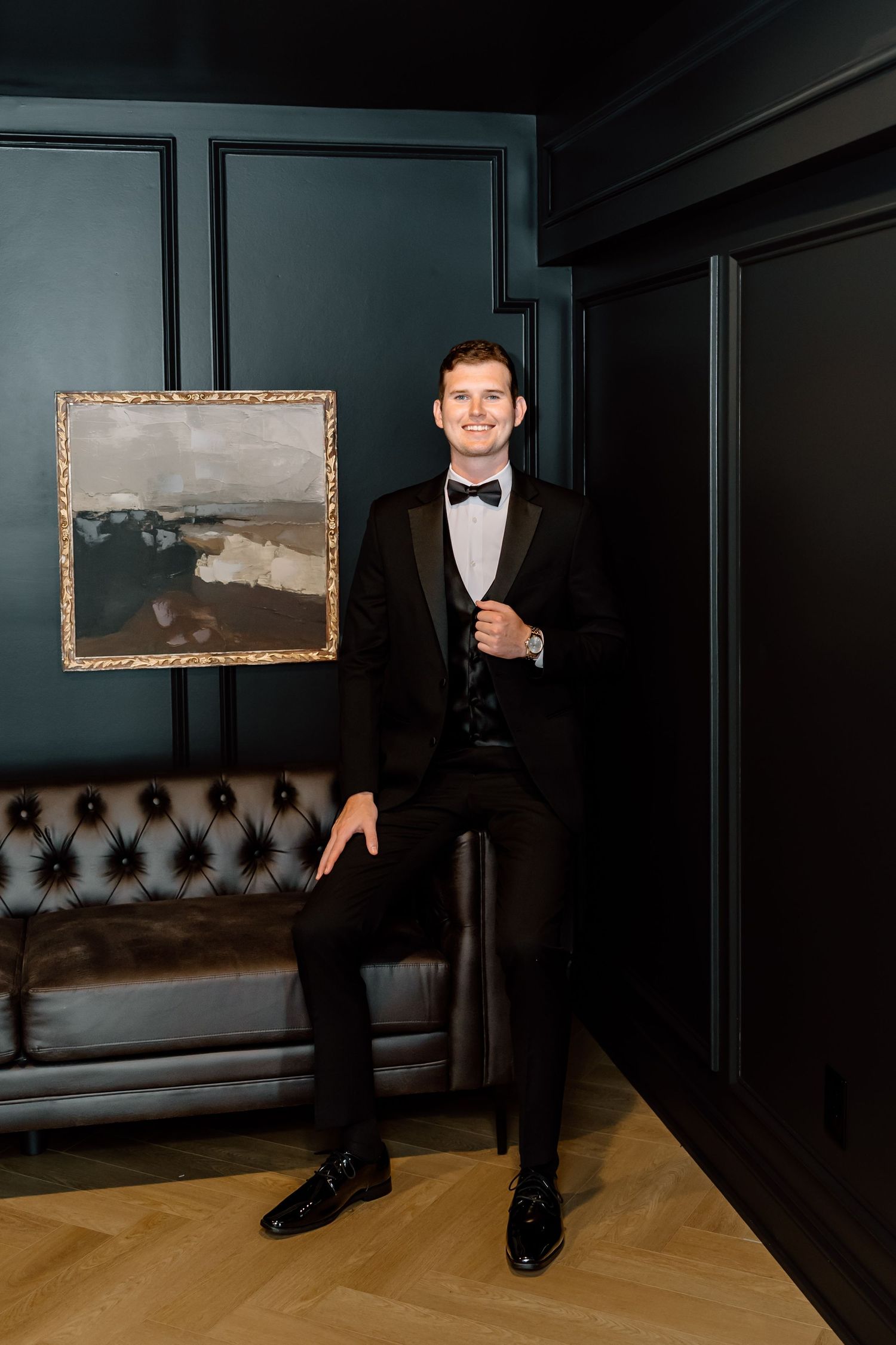 A smiling man in a black tuxedo with bow tie poses confidently in an elegant dark-paneled room with leather sofa.