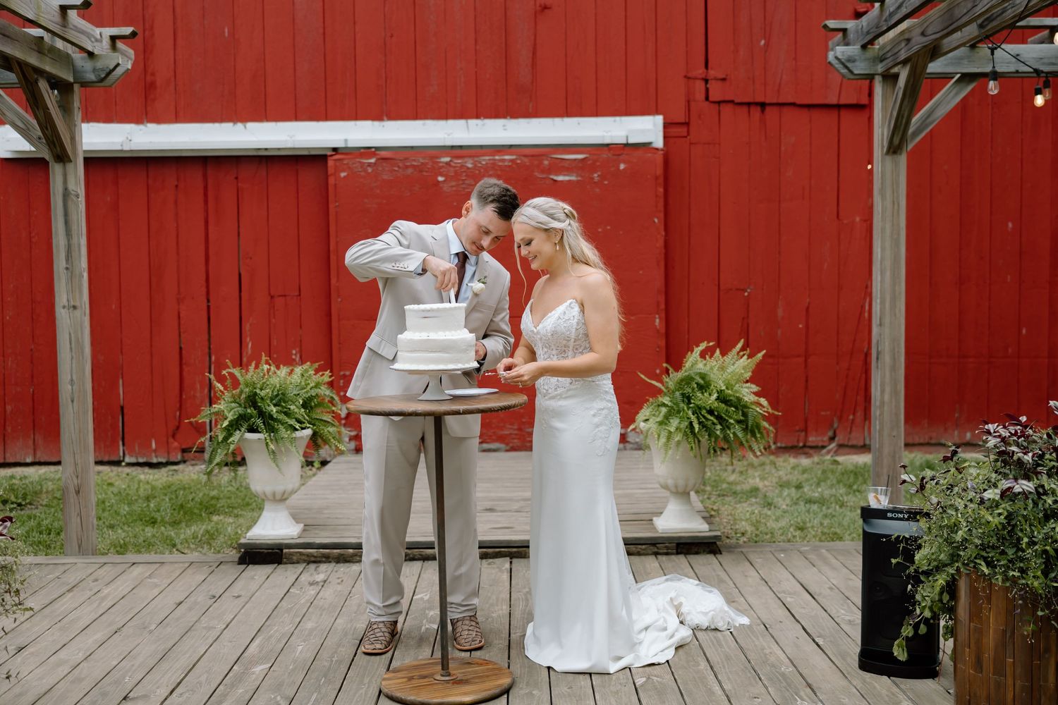 Bride and groom cutting wedding cake on rustic wooden deck in front of red barn.