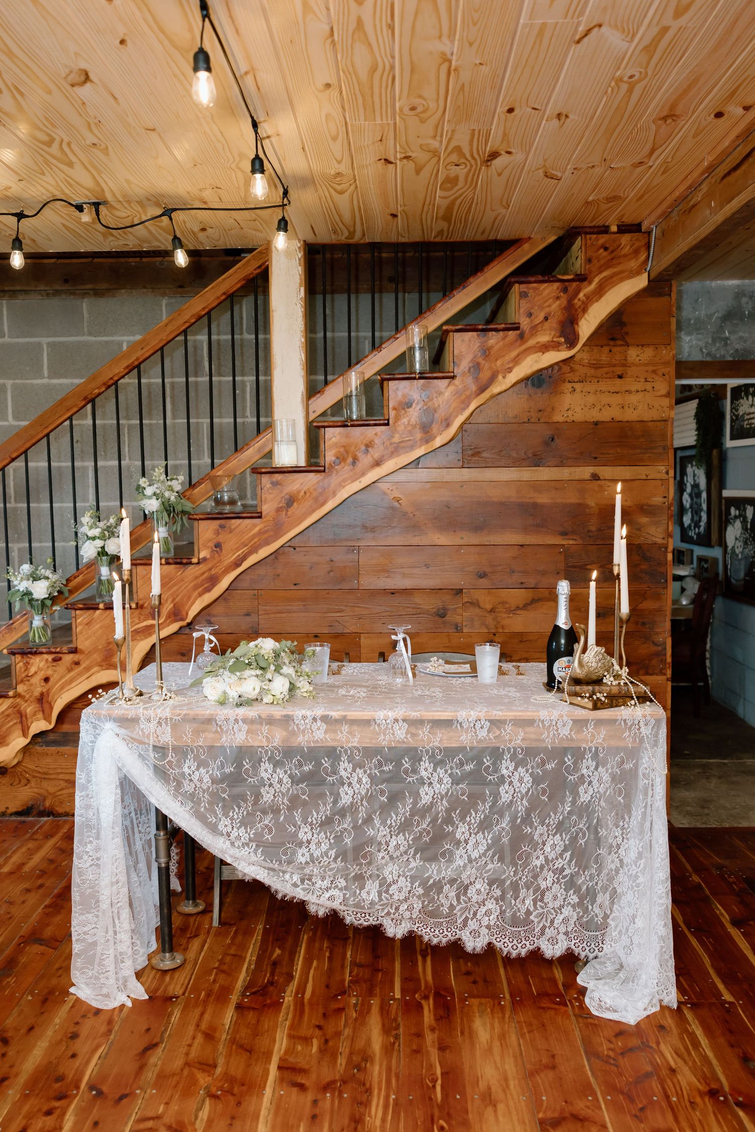 Rustic wedding sweetheart table with lace tablecloth, candles, and floral decor beneath a wooden staircase.
