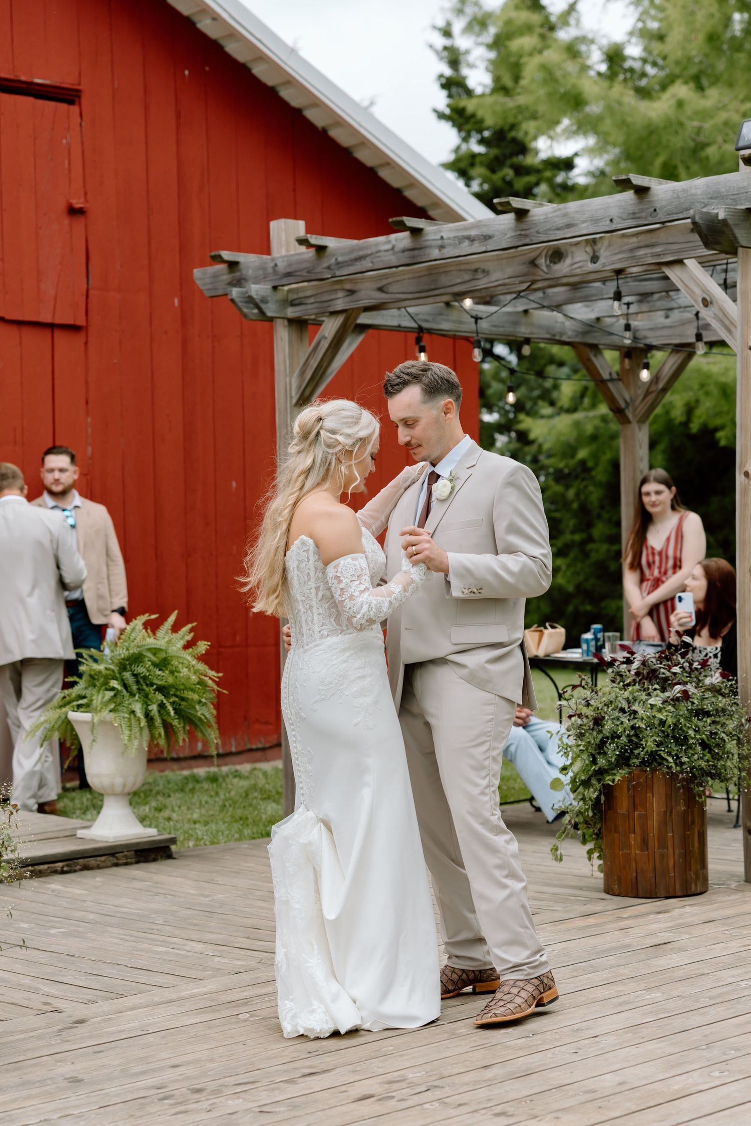 Bride and groom share first dance on rustic wooden deck beside a red barn at outdoor wedding.
