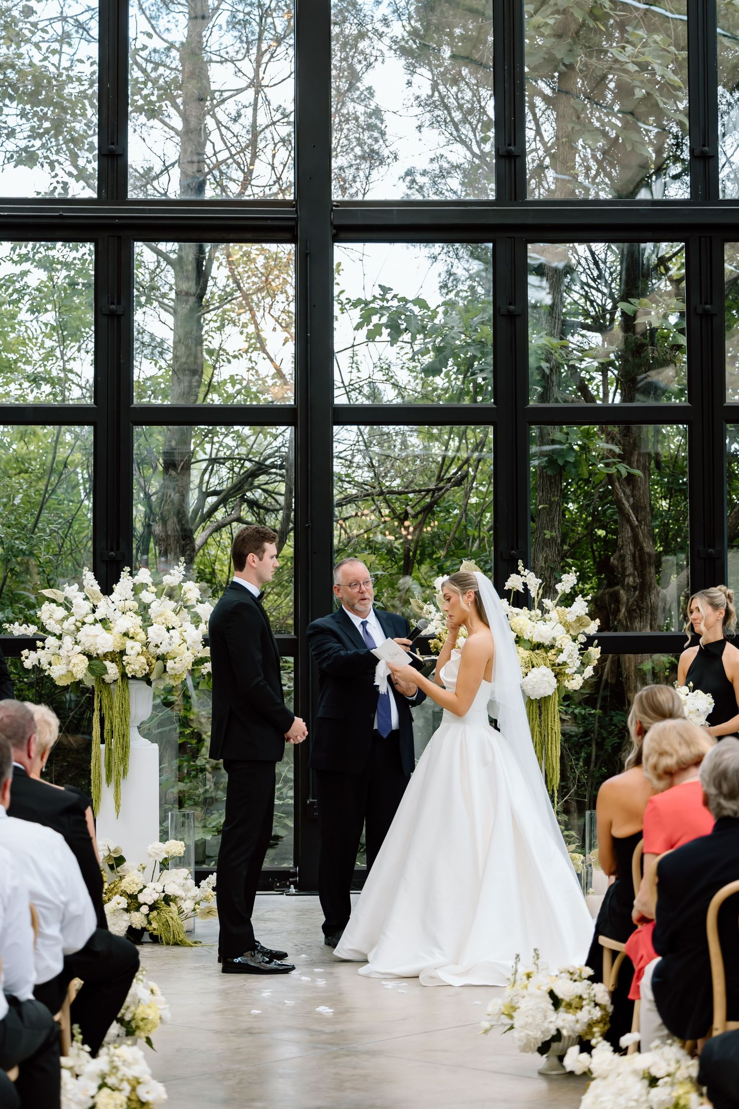 Bride and groom exchange vows at elegant indoor wedding ceremony with white floral arrangements and floor-to-ceiling windows.