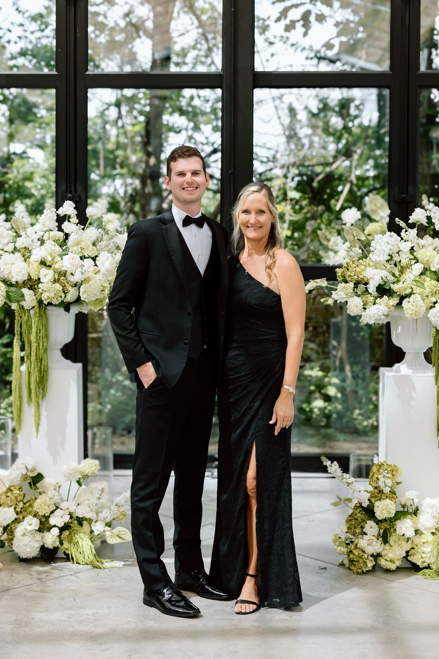 A man in a black tuxedo and woman in a black gown pose together among white floral arrangements at a formal event.