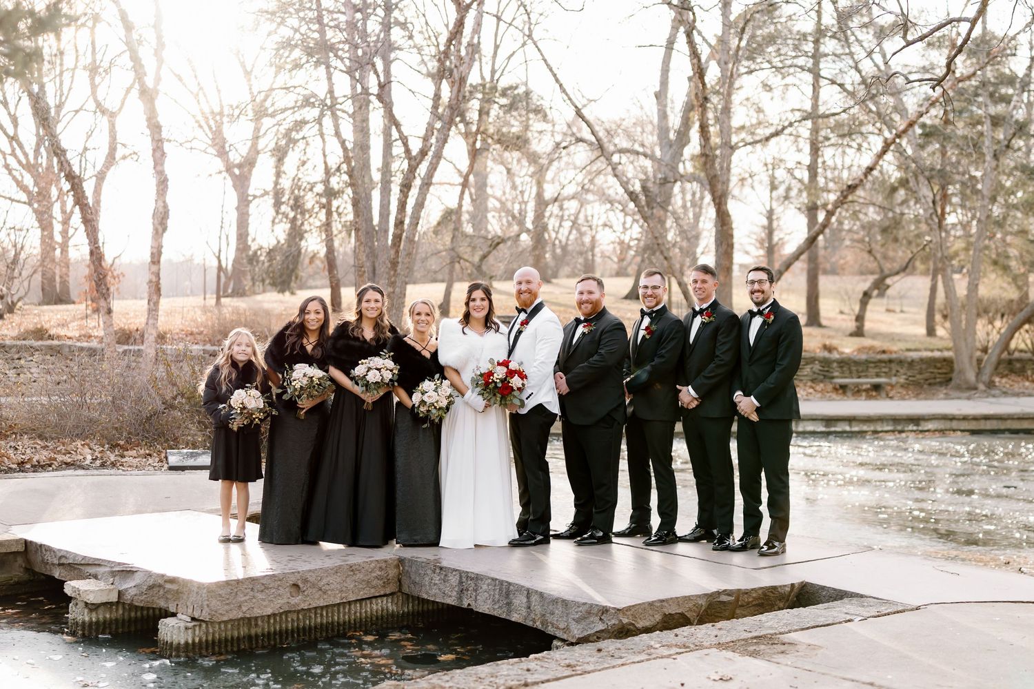 A wedding party in black and white attire poses outdoors in a snowy winter landscape with bare trees in the background.