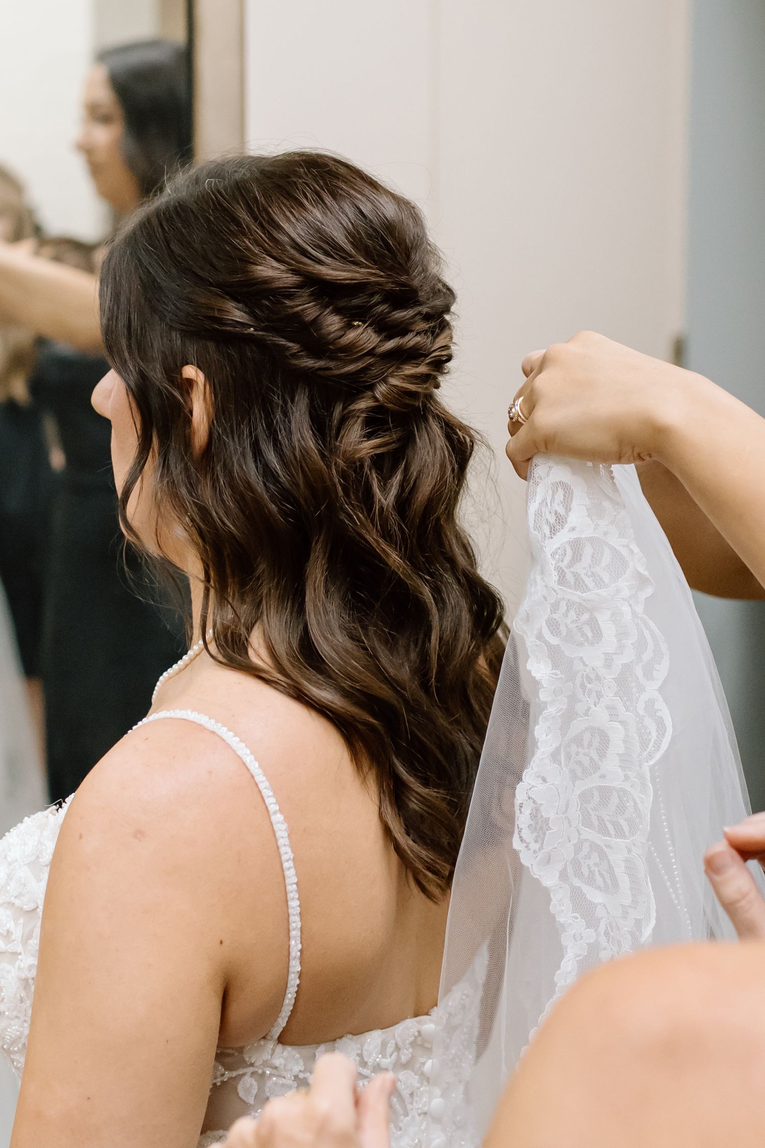 A hairstylist adjusts a delicate lace veil while styling an elegant braided half-up wedding hairstyle.