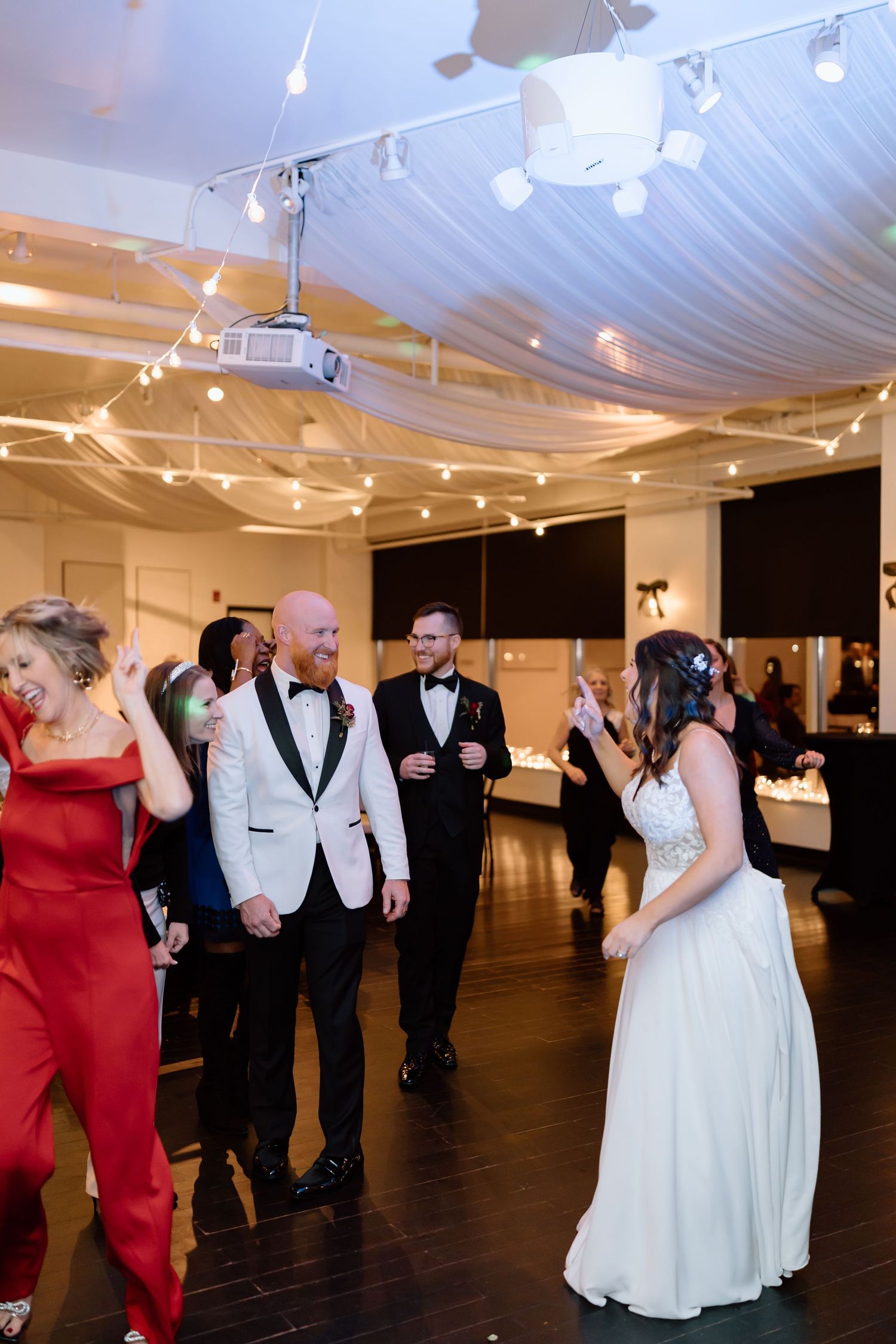 Wedding guests dance together under white tent lighting at an indoor reception venue.