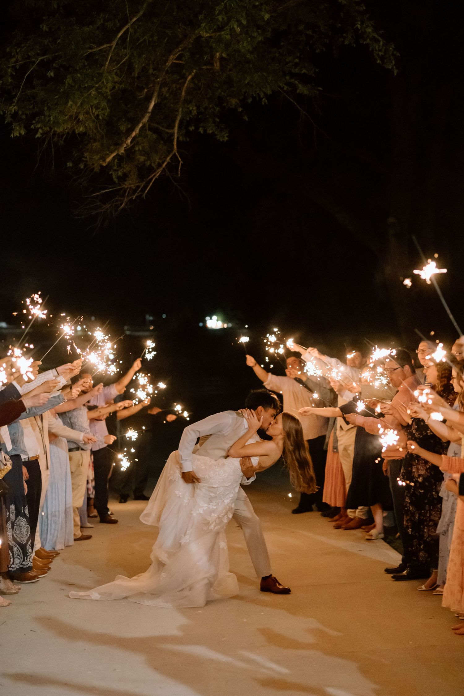 Bride and groom share a romantic dip kiss surrounded by guests holding sparklers at an outdoor nighttime wedding.
