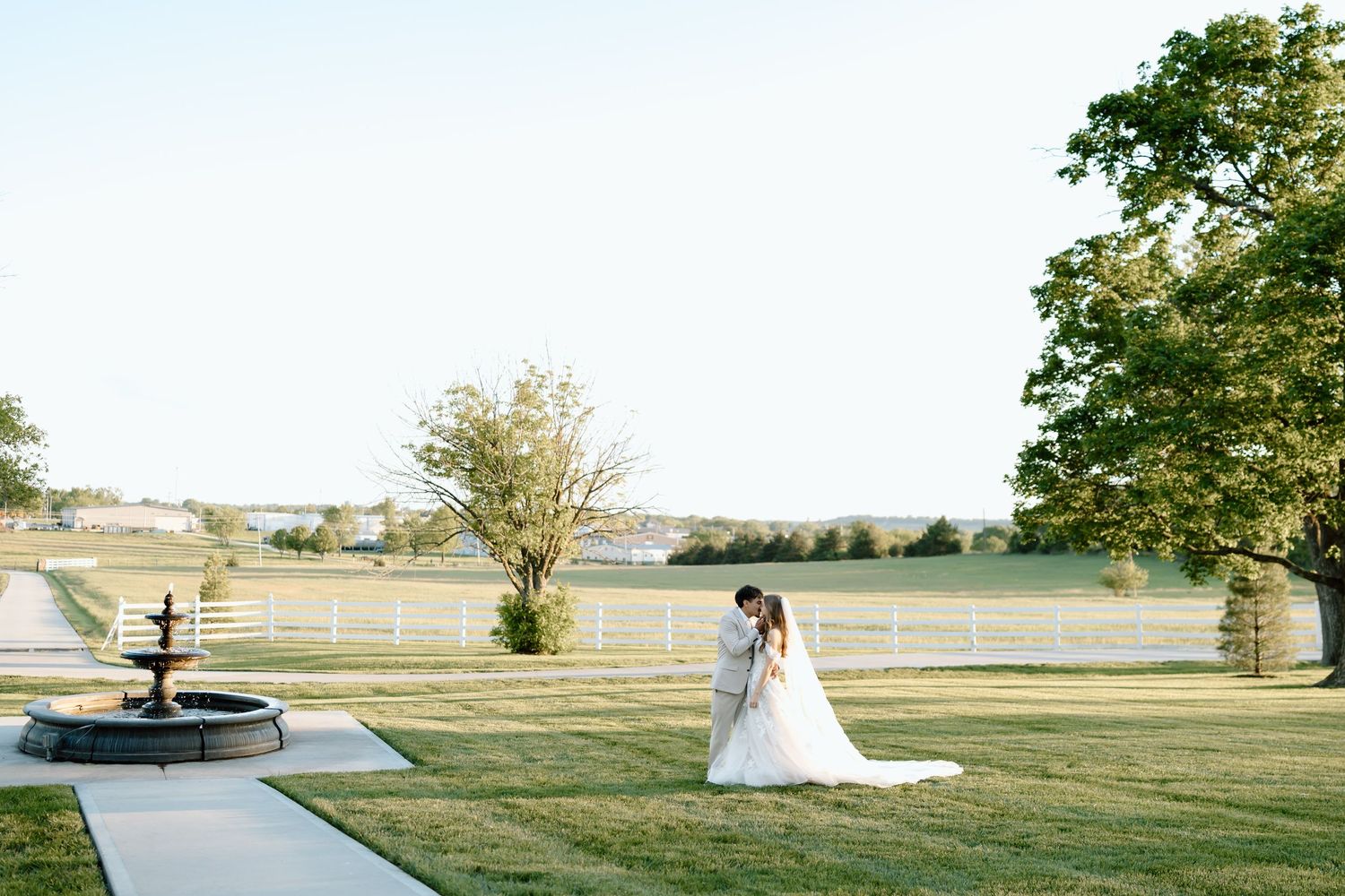Bride and groom share a romantic moment on lush green lawn beside a fountain at an elegant outdoor wedding venue.