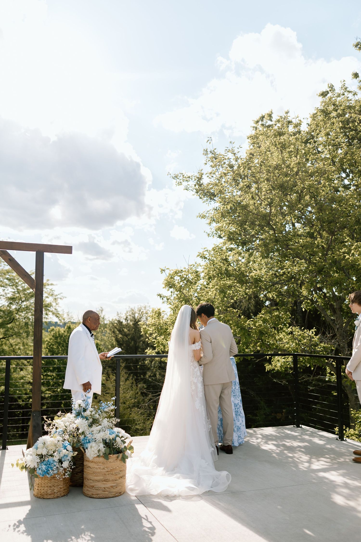 Outdoor wedding ceremony on a sunny terrace with bride in white gown, groom in tan suit, and officiant reading vows.