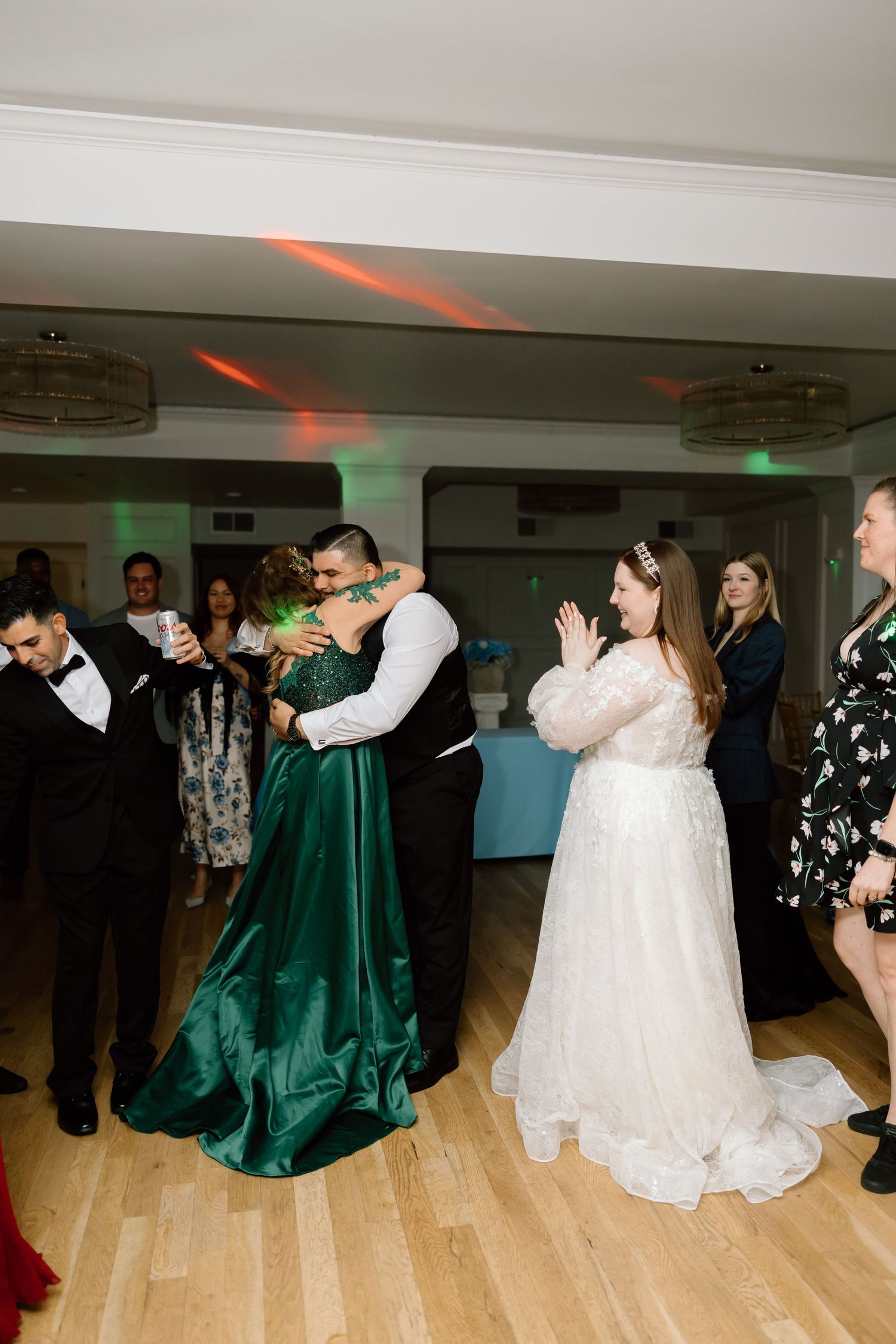 Bride in white gown watches as groom embraces woman in emerald green dress at wedding reception dance floor.