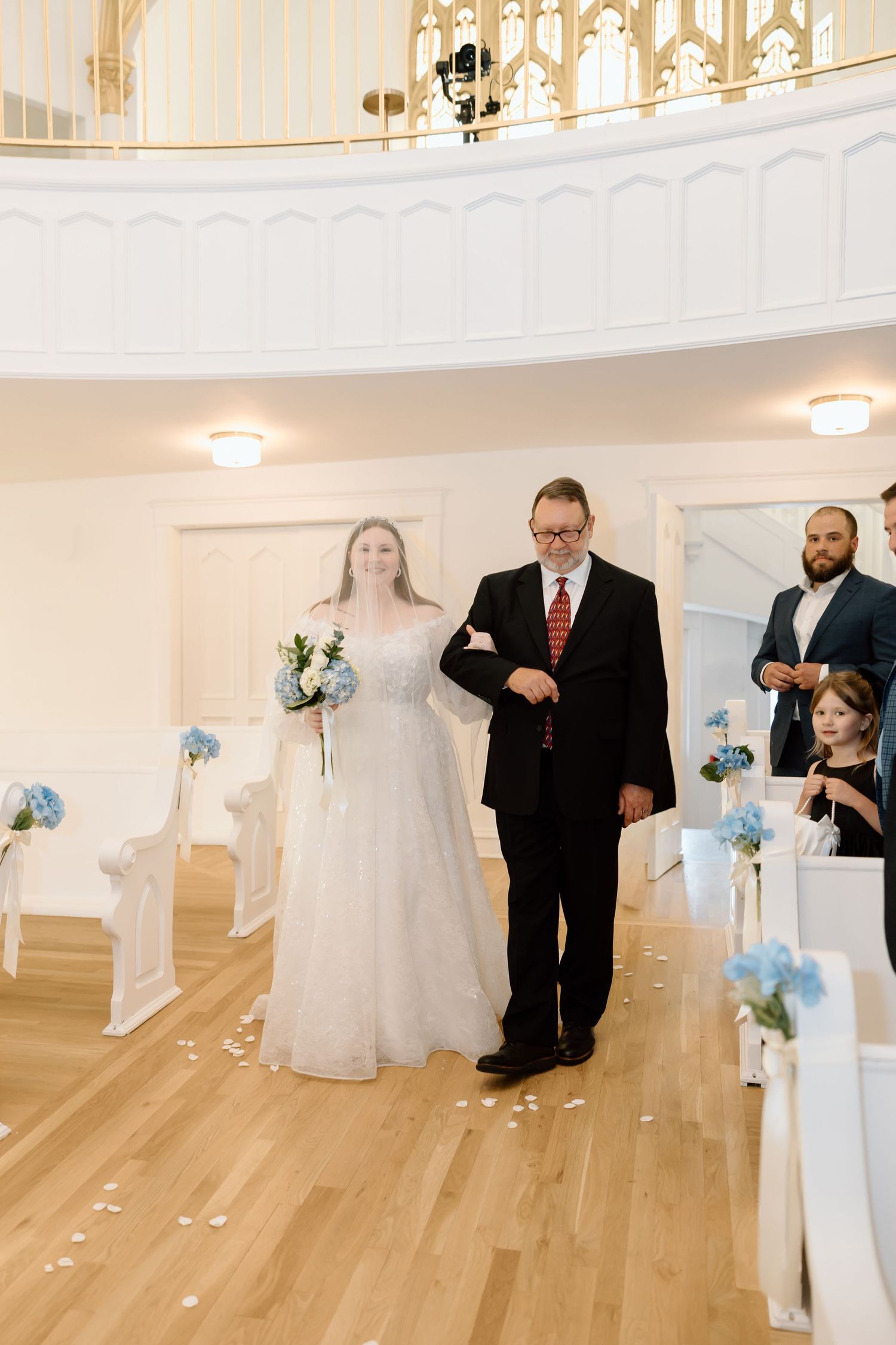 Bride in white gown walks down aisle with escort in black suit in elegant white wedding chapel.
